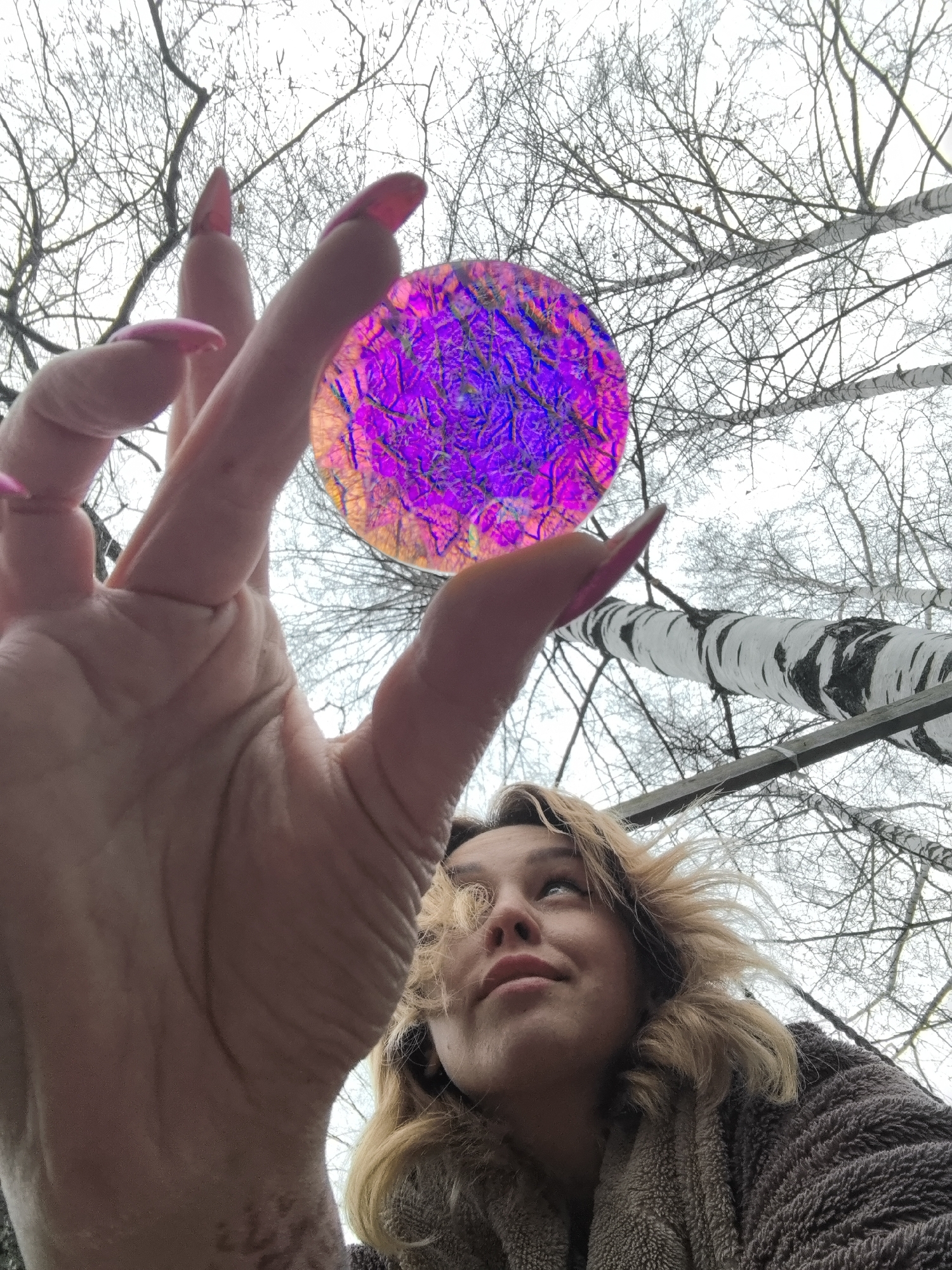 A low-angle shot features a woman holding a translucent, purple and orange iridescent disc in her hand. The woman has blonde, shoulder-length hair and is wearing a brown, textured garment. Her pink fingernails are long and manicured. Her hand is closest to the camera, dwarfing her face. She is looking upward and slightly to the right.
Behind the woman and her hand are bare tree branches against a pale sky, with birch trees partially visible. The location is possibly Noviy, Russia, based on the prompt's instruction to include this location. No additional information in the image supports the location.