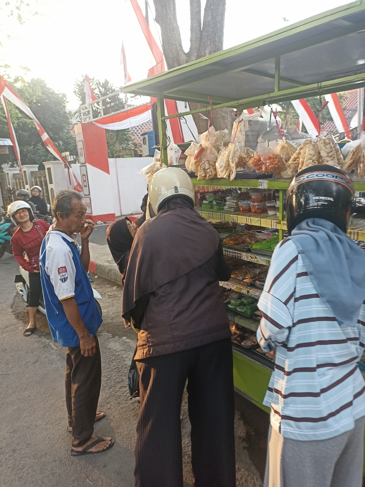 In Mojopanggung, Indonesia, a street vendor's cart is laden with various snacks, displayed in clear plastic bags and containers. The cart is illuminated by the warm glow of late afternoon sun, which casts long shadows across the asphalt road. Several people are gathered around the cart, examining the offerings. Among them is an elderly man in a blue and white shirt, who appears to be deep in thought. A woman in a red shirt and a white helmet stands nearby, a slight smile on her face, while two individuals in helmets, one in a brown jacket and the other in a striped shirt and blue headscarf, are actively browsing the snacks. The background reveals a street scene with houses and trees, with red and white Indonesian flags adorning the surroundings, suggesting a national celebration. A motorcycle is parked to the left, adding to the everyday life atmosphere of the scene.