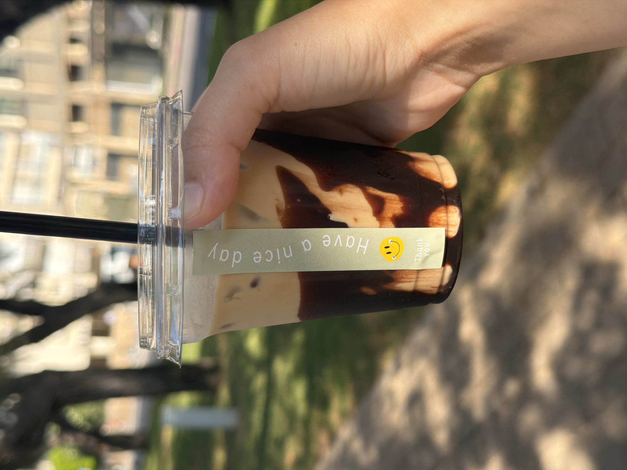 A person's hand holds a clear plastic cup containing an iced coffee drink with a visible layer of chocolate syrup. A light-colored sticker is affixed to the cup, featuring the text "Have a nice day" in a vertical orientation, accompanied by a smiling face emoji and the words "Thank you." The cup is presented outdoors, with a blurred background suggesting a city environment in Bat Yam, Israel. The background includes trees, paved ground, and buildings. The lighting indicates it is daytime, with sunlight casting shadows.
