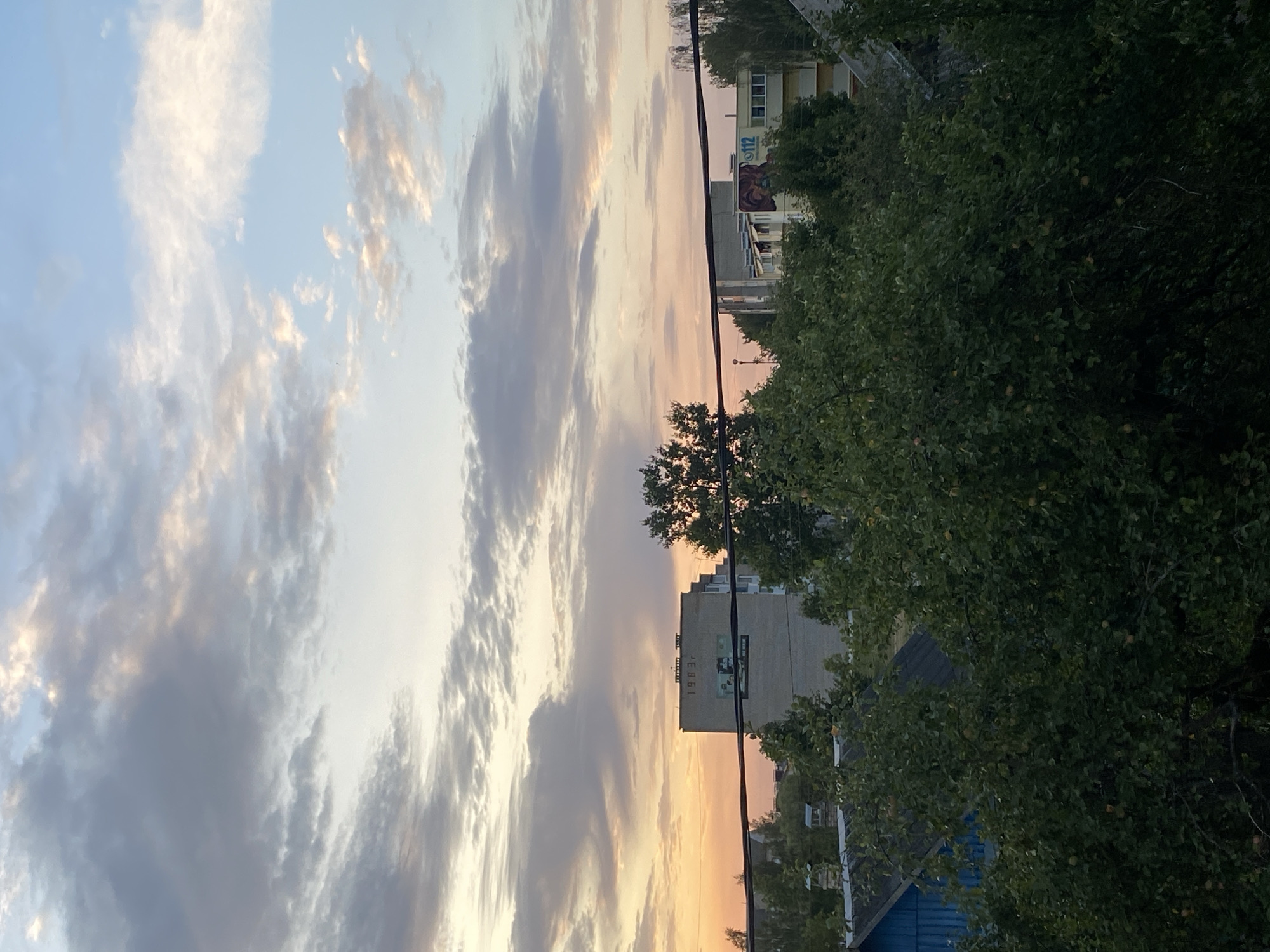The image depicts a view of buildings and trees against a twilight sky. In the foreground, dense green foliage from trees partially obscures the scene. Behind the trees, a series of low-rise structures and a taller, rectangular building are visible. The taller building has a mural or large graphic on its facade and the year "1983" is marked near its top. A dark, horizontal power line stretches across the middle of the frame. The sky above is a gradient of pale orange and pink near the horizon, transitioning to light blue and gray clouds higher up. The image appears to be taken from an elevated viewpoint, looking out over a residential or mixed-use area within the city of Krupki, Belarus.