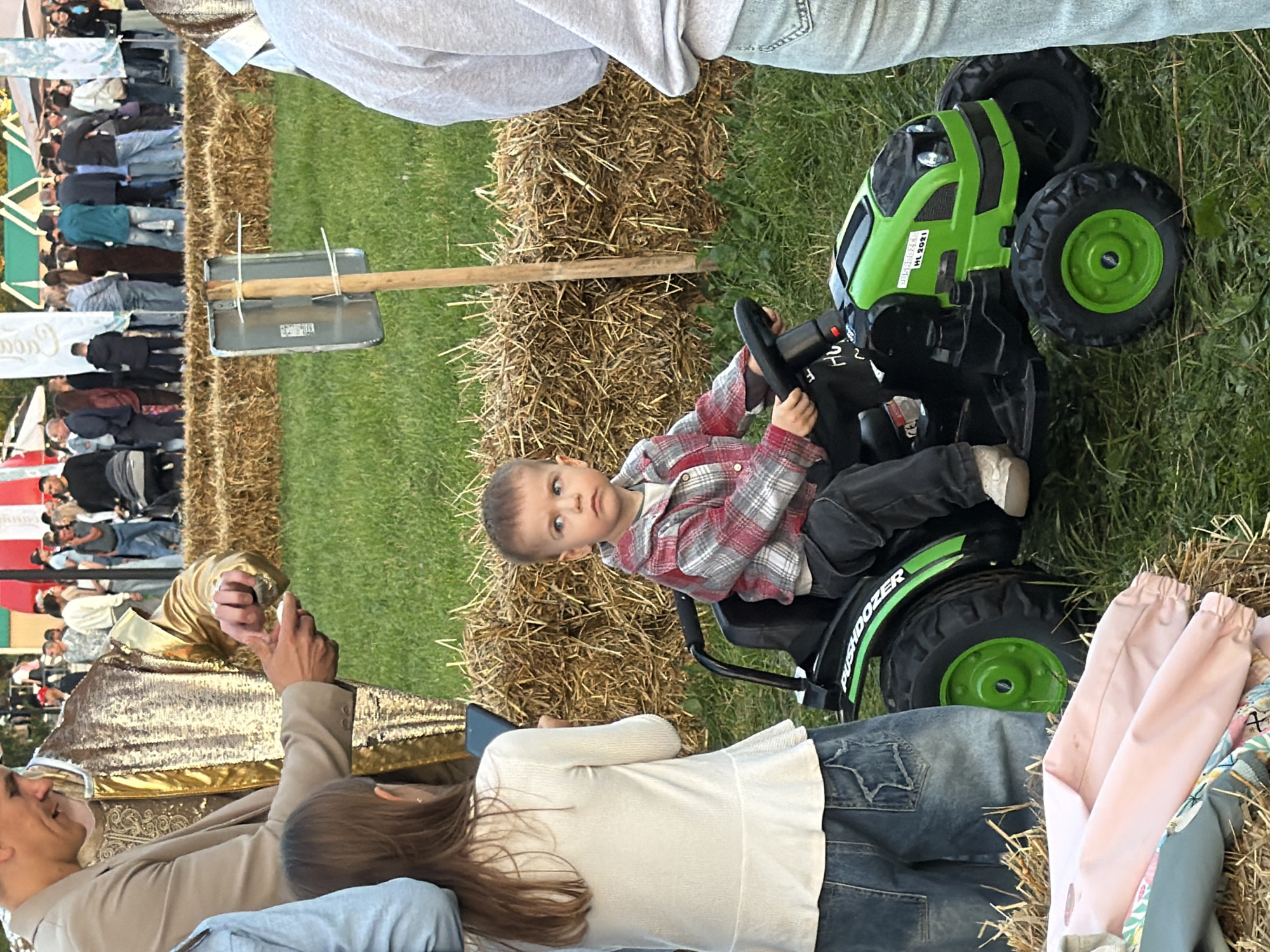 In a grassy area with hay bales, a young boy, wearing a red and gray plaid shirt and dark pants, sits at the steering wheel of a green toy tractor. He looks to his right, with his hands on the wheel. To his left, a young girl with long brown hair, dressed in a white long-sleeved shirt and blue jeans, stands holding a dark rectangular object, possibly a phone. Behind the children, a person in a tan suit with gold sequined accents is visible, appearing to interact with something in their hands. In the background, a crowd of people are gathered, some standing behind hay bales, with tents and banners visible. The scene appears to be an outdoor event.