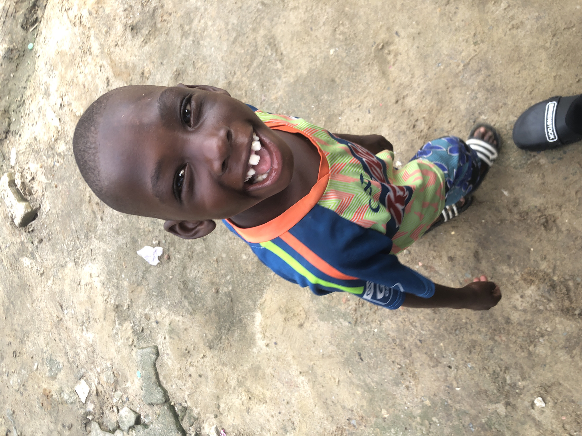 A young Black boy, with a bright smile and missing front teeth, stands in an outdoor, dirt-covered area. He is wearing a blue and orange t-shirt with green and white stripes and blue shorts with a floral pattern. He has on black and white sandals on his feet, and another black sandal is visible on the ground to his right. The background consists of rough, uneven ground with scattered debris, suggesting an unpaved area. The image appears to have been taken from a slightly elevated perspective. The scene is consistent with an outdoor setting in a neighborhood in Ado, Nigeria.