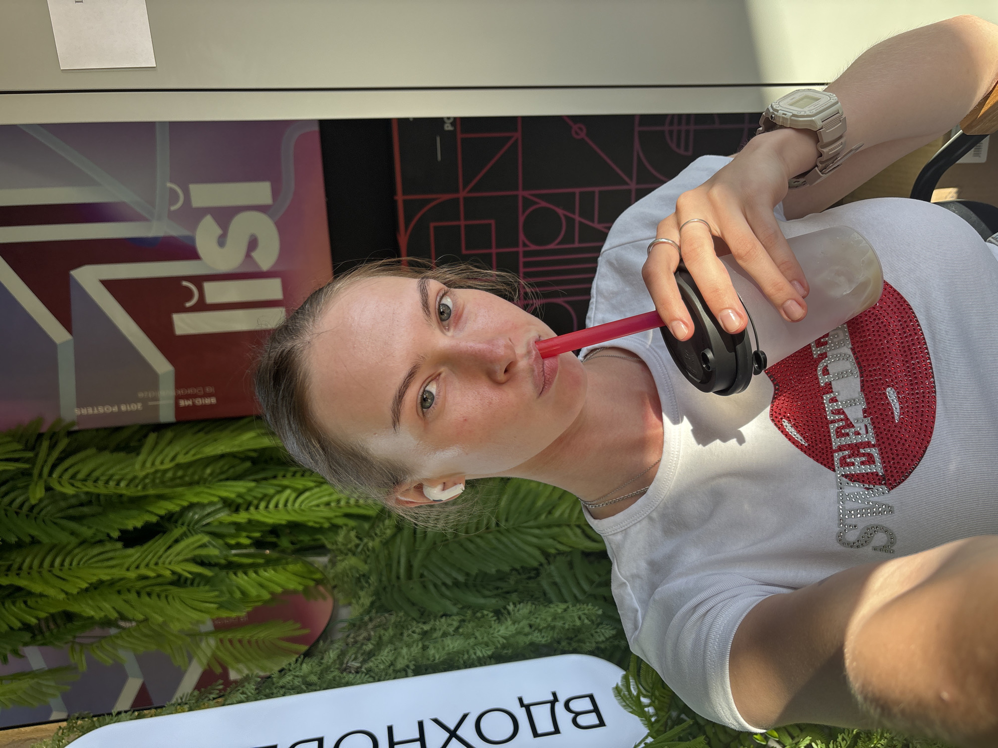 A young woman with light skin and blonde hair pulled back is taking a selfie while drinking from a translucent cup with a red straw. She is wearing a white t-shirt with a red sequined image of lips and the word "SWEET" spelled out in rhinestones. She has a silver necklace and several rings on her fingers, as well as a light-colored watch on her left wrist. She is also wearing white wireless earbuds. She is outdoors, with lush green foliage to her left and a wall with a geometric design and text to her right. The text on the wall appears to be in Cyrillic, with "l isi" prominently displayed. The text on a white banner on the left side of the image is also in Cyrillic and reads "ВДОХНОВ". The scene appears to be an outdoor seating area of a cafe or restaurant in Perm, Russia, possibly a trendy establishment given the decor. The lighting suggests it is daytime, with sunlight casting shadows.
