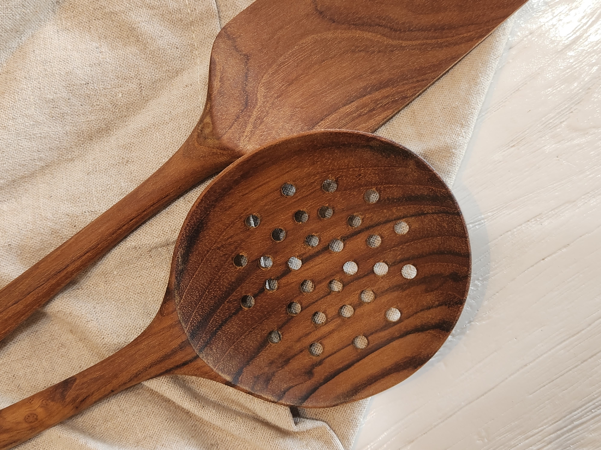Two wooden cooking utensils, a slotted spoon and a spatula, lie on a piece of light-colored, woven fabric. The slotted spoon, made of dark wood with visible grain, is positioned in the foreground and angled to the left. It features numerous circular perforations across its surface. The spatula, also made of wood with a similar grain pattern, is placed behind and to the right of the spoon, its flat, angled head partially visible. The fabric is laid out on a white, horizontally-lined surface, possibly a table or countertop. The image is a close-up shot, with the lighting casting soft shadows and highlighting the textures of the wood and fabric. The scene appears to be indoors, with no discernible city or geographical context from Maitland, United States of America.