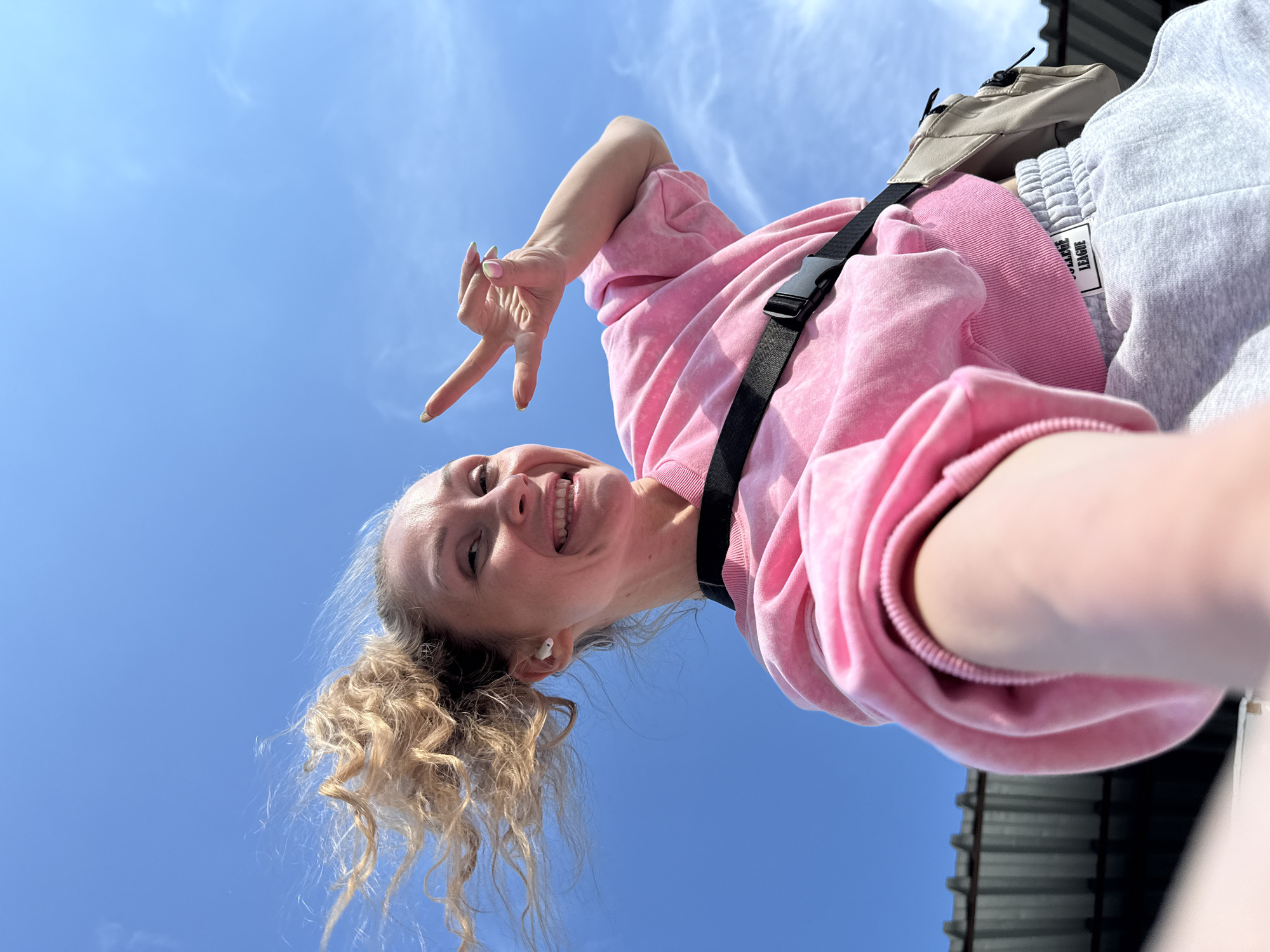 A young woman with blonde, curly hair pulled back in a ponytail is looking up at the camera, smiling. She is wearing a pink tie-dye sweatshirt and grey sweatpants with a label that reads "COLLEGE LEAGUE". A black strap, part of a crossbody bag, is visible across her chest. She is holding up her right hand, with her index and middle fingers extended in a peace sign, and her thumb pressed against her ring finger. Her left hand is partially visible in the lower right corner of the frame, appearing to hold the camera. The background is a clear blue sky with a few wispy white clouds. The camera is positioned at a low angle, looking up at the woman. The sunlight suggests it is daytime. The overall mood is cheerful and energetic.