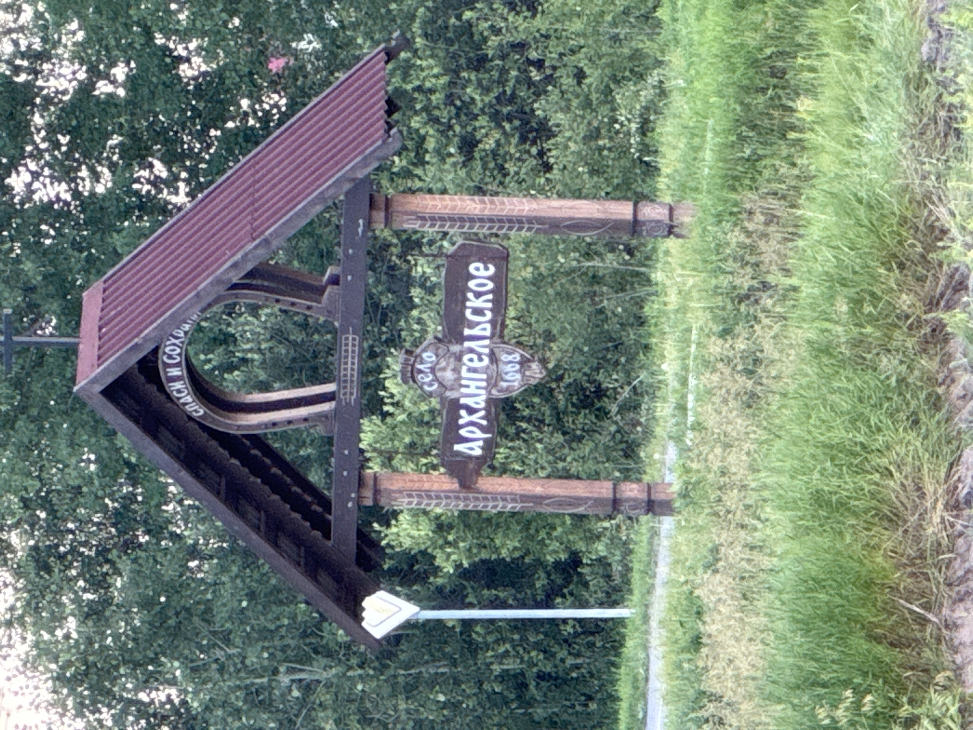 The image shows a wooden signpost in the village of Arkhangelskoye, Russia. The signpost is located in a grassy area with trees in the background. The sign itself is brown with white lettering and depicts the name of the village, "Архангельское," and the year "1668." Above the village name, there is text that reads "СПАСИ И СОХРАНИ," which translates to "Save and Protect." The signpost is supported by two wooden posts carved with decorative patterns. A white diamond-shaped sign is visible on a pole to the left of the main signpost. The overall scene suggests a rural or historical setting.