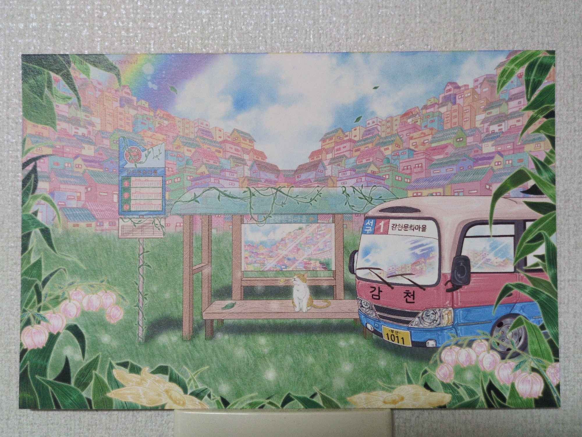 The image is a postcard of an illustration depicting a bus stop in a colorful, hillside town. The town is composed of closely packed, pastel-colored houses. A rainbow is visible in the sky above the town. In the foreground, a bus stop is constructed of wood with vines growing on it. A white and orange cat sits on the bus stop bench. To the right is a pink and blue bus with “감천” written on it in Korean. The bus license plate reads “부산 1011”. A bus route marker reads "석 1 감천문화마을". Flowers are depicted growing in the foreground.