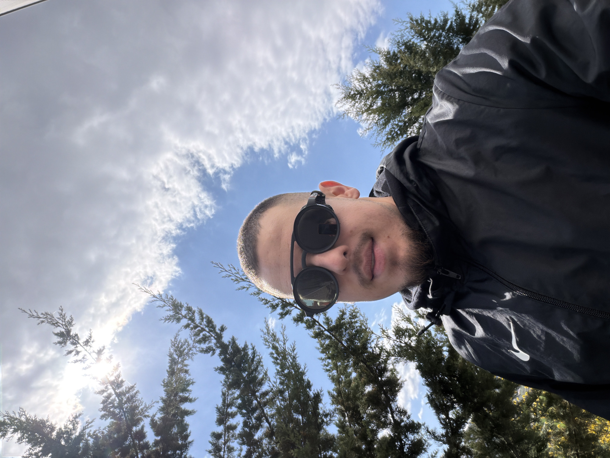 A medium-shot, angled upward, depicting a man wearing black sunglasses and a black hooded jacket, possibly a Nike jacket. He has short-cropped hair and a beard. Behind him is a sky with white clouds and tall, green evergreen trees. The location is potentially Parvenets, Bulgaria.