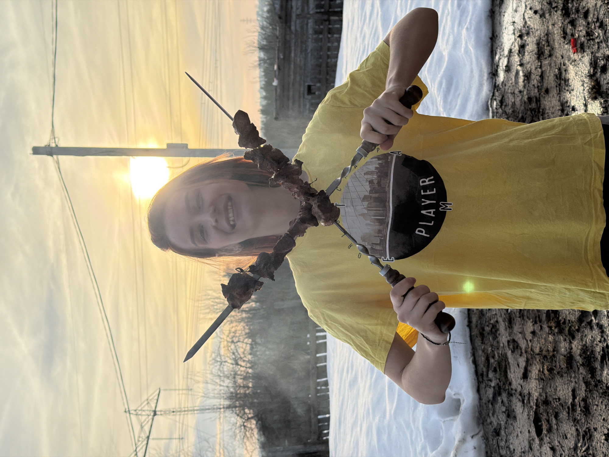 The image depicts an outdoor scene in Chaikovsky, Russia, at sunset or sunrise, characterized by low, warm sunlight illuminating the background. A young individual, appearing male, with reddish-brown hair, is positioned centrally, smiling directly at the camera. They are wearing a bright yellow short-sleeved t-shirt featuring a dark circular graphic on the chest with a cityscape silhouette and the text "PLAYER M". A dark beaded bracelet is visible on their left wrist.
The individual holds two metal shashlik skewers horizontally, crossed in an 'X' shape in front of their chest. Each skewer is laden with several pieces of cooked, dark meat. Faint smoke is visible rising from or immediately behind the skewers on the left side.
The foreground consists of dark, muddy soil mixed with patches of melting snow. Further back, a significant bank of white snow extends across the middle ground. In the far background, bare deciduous trees and bushes are visible. Utility infrastructure includes a tall metal electricity pylon on the left and a wooden utility pole on the right, both strung with power lines. A faint, dark wooden structure is visible behind the snow on the right side. The sky displays a clear horizon with a strong sunburst on the right, transitioning to a pale, cloudy upper sky. A small, red object lies on the muddy ground in the bottom right corner.