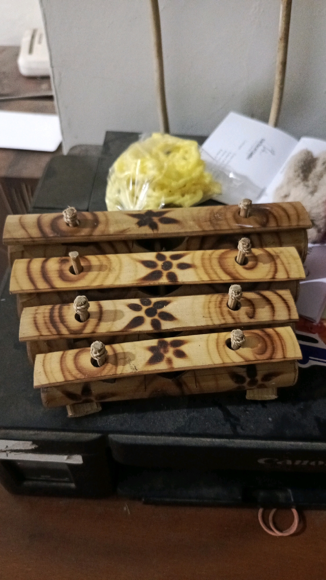 The image shows a wooden xylophone on a black surface, possibly a printer. The xylophone has four wooden bars, each decorated with a floral pattern burned into the wood. Each bar has a small wooden peg inserted through a hole. In the background, a white wall and a bundle of yellow material wrapped in plastic are visible. The scene is lit by ambient light. The objects are arranged in a still life composition.