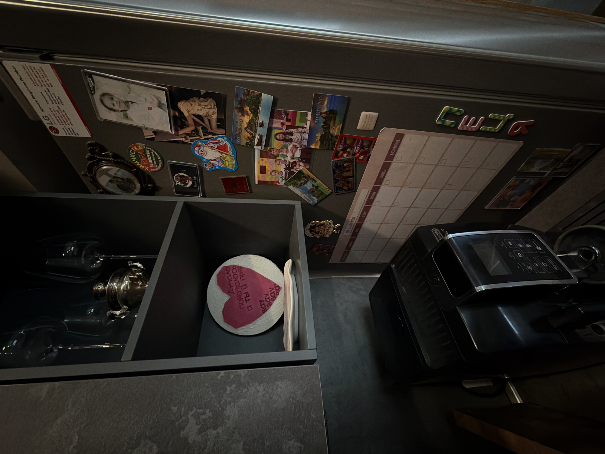 The image shows a close-up view inside a kitchen. A refrigerator door covered with various magnets and images is visible. Magnets include vacation souvenirs labeled "THAILAND," a calendar, an LG Service telephone number, and a handwritten paper that says “Thought it seemed, and you are even straight.” A gray cabinet with two shelves sits to the left. The upper shelf contains drinking glasses and a metal tea set, and the lower shelf holds a heart-shaped decorative item. To the right of the cabinet and against the refrigerator door is a black DeLonghi coffee machine. The image suggests an indoor setting within a private residence, likely in a kitchen. There is no indication that the photo was taken in Birobidzhan, Russia.