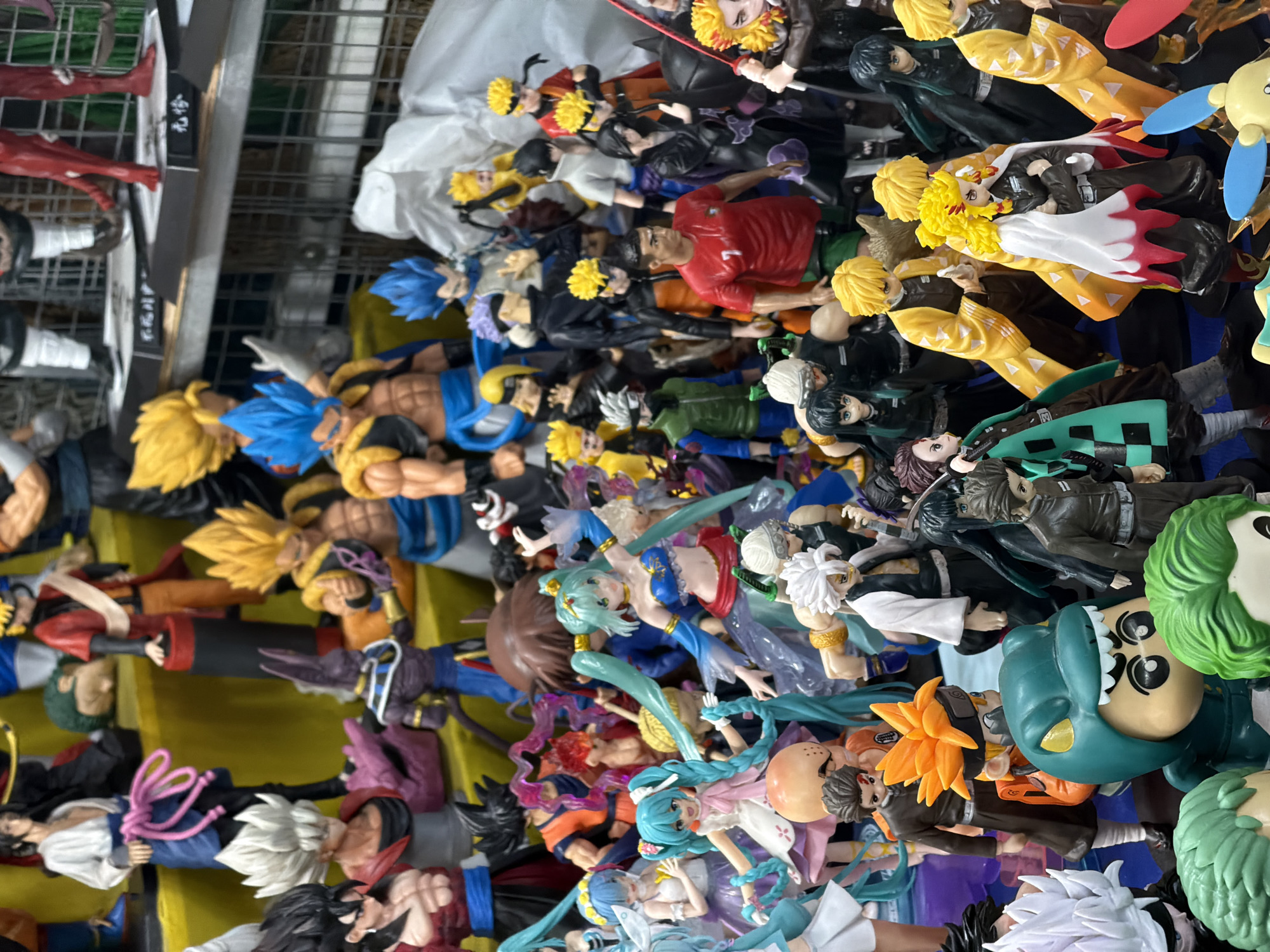 The image shows a dense collection of anime figurines. The figurines are diverse, featuring characters with vibrant hair colors like yellow, blue, and green. There are Dragon Ball Z, Demon Slayer, and Hatsune Miku characters. Some figurines appear to be duplicates. The figurines are tightly packed, and the background shows a wire shelving unit. The location of the image is in Da Nang, Vietnam.