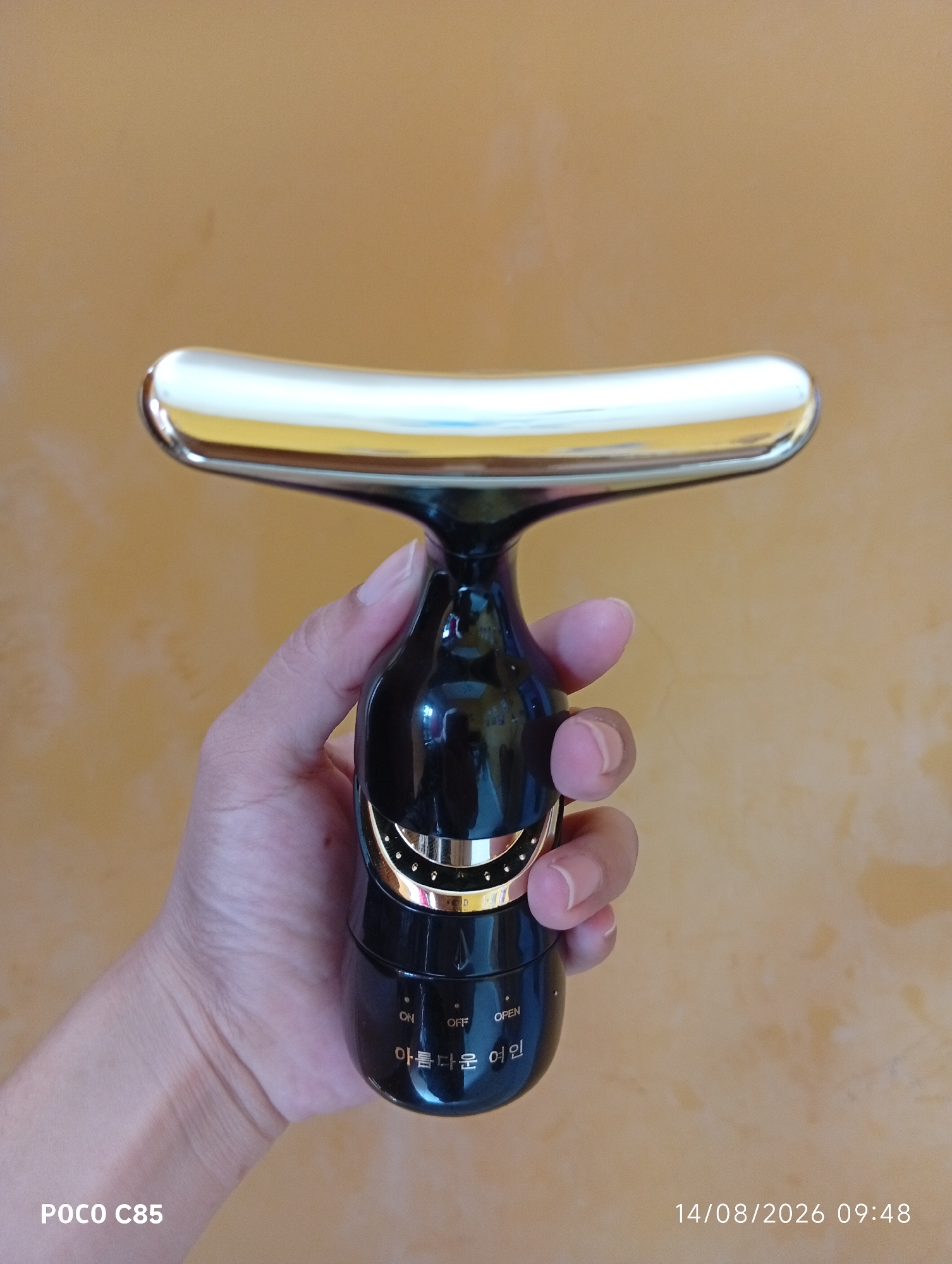 A close-up, high-angle shot shows a hand holding a black and gold electric facial roller against an orange, textured background. The roller has a T-shaped head with a white and gold curved surface and a black, curved handle with gold accents. On the handle, there are markings that appear to be "ON", "OFF", and "OPEN", along with Korean text. The hand's fingers are wrapped around the handle. The bottom left corner of the image displays "POCO C85", and the bottom right corner shows the date "14/08/2026 09:48". The setting appears to be indoors, likely a home environment, and there are no visible elements to suggest it is in South Tangerang, Indonesia.