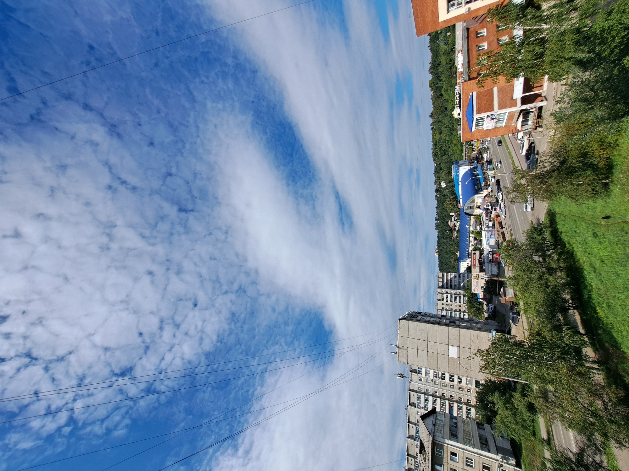 The image shows a view of an urban area with apartment buildings, commercial structures, a road, and trees under a partly cloudy sky. The architecture of the buildings, particularly the multi-story apartment blocks with balconies and the brick commercial buildings, suggests a post-Soviet era development common in many Russian cities. The presence of Cyrillic signage on the commercial buildings confirms this. The style of the apartment buildings, with their concrete facades and prominent balconies, is characteristic of the Soviet modernist architectural style prevalent in the late 20th century. The road appears to be a main thoroughfare with several cars driving on it. The surrounding greenery indicates a temperate climate. The overall impression is that of a typical residential and commercial district within a Russian city.