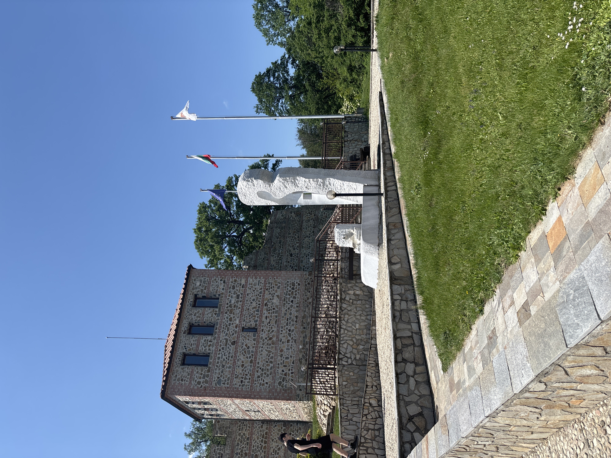 The image depicts a terraced historical site on a clear, sunny day in Belchin, Bulgaria, specifically the Tzari Mali Grad fortress. In the center-left, a multi-story stone and brick building, likely a reconstructed medieval tower, stands with a reddish-tiled roof from which a tall, thin antenna or lightning rod extends vertically into the sky. Its facade features several rectangular windows with dark frames.
In the mid-ground, to the right of the building, a large, abstract white stone monument with an elongated, hollowed form is positioned on a paved platform. A smaller white stone structure rests at its base. Three flagpoles rise behind the monument: from left to right, they fly the blue and yellow-starred flag of the European Union, the white-green-red horizontal tricolor flag of Bulgaria, and a white flag bearing a red symbol, possibly a local crest.
A male individual, dressed in a dark t-shirt, dark shorts, and dark shoes, walks uphill on a cobblestone pathway in the lower-left quadrant, moving towards the building. He is mid-stride, with his left leg forward.
The site features multiple levels of terraced landscaping with retaining walls constructed from irregular natural stones. Paved walkways are made from various stone slabs and cobblestones. Dark metal railings with intermittent support posts are present along the walkways and stairs. Two spherical, dark-colored ground lamps are visible near the white monument. The foreground consists of vibrant green grass with scattered small white flowers. Lush green deciduous trees border the scene in the background and to the sides. The sky is a uniform, clear blue, indicating bright, daylight conditions.