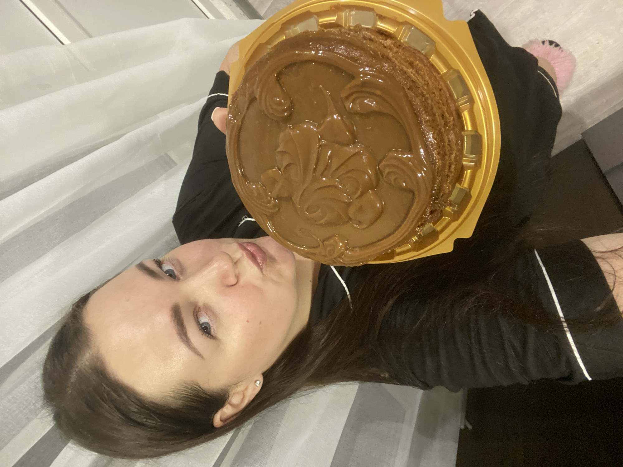 The image, presented sideways, shows a young woman lying down indoors. She has dark brown hair, blue eyes, and fair skin, wearing a black long-sleeved top with white piping and a small stud earring. She is holding a round, chocolate or caramel-colored cake in a golden plastic container up towards the camera. The cake is adorned with a glossy, swirled frosting design, featuring an ornate, possibly stylized fleur-de-lis pattern in the center. Distinct layers are visible along the cake's sides. The woman's expression is neutral and attentive, looking towards the cake or the viewer. The background consists of white and grey striped fabric, likely bedding or a large curtain. A pink fluffy slipper is visible on her right foot. The indoor setting and soft lighting suggest it might be evening or night. No text is visible in the image. The location context provided for the scene is Sochi, Russia.