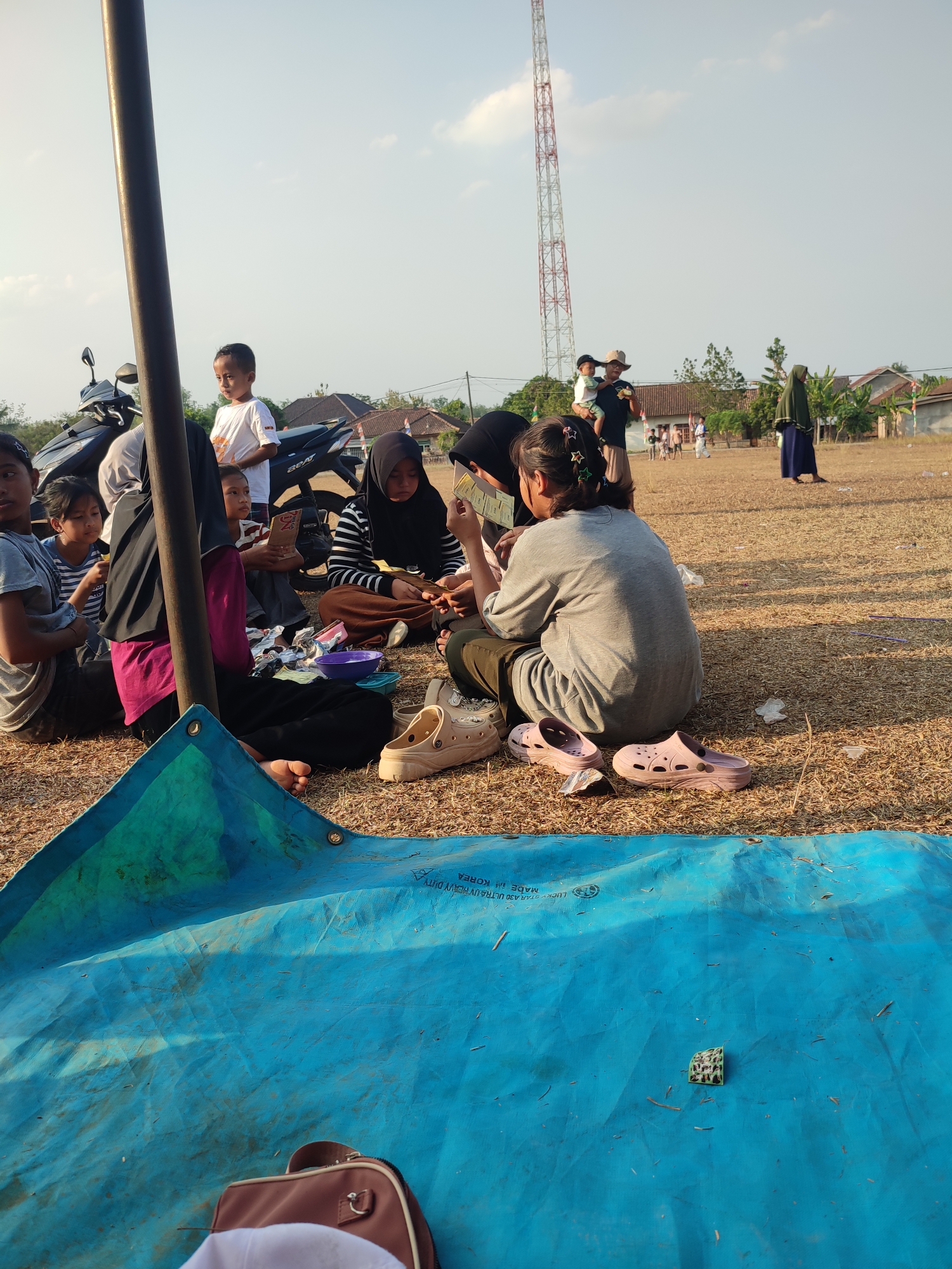 In Nusa Jaya, Indonesia, a group of people are gathered outdoors in a dry, grassy field. The scene is illuminated by natural sunlight, suggesting it is daytime. In the foreground, a large blue tarp is spread out, partially covering a brown bag and a white cap. Surrounding the tarp, several individuals are seated on the ground, some appearing to be engaged in conversation or sharing food. A child in a white t-shirt stands near a parked motorcycle. In the background, a red and white telecommunications tower is visible, along with houses and trees, indicating a semi-rural or suburban environment. Several other people are scattered across the field, some walking and others standing. The overall atmosphere suggests a casual gathering or picnic.