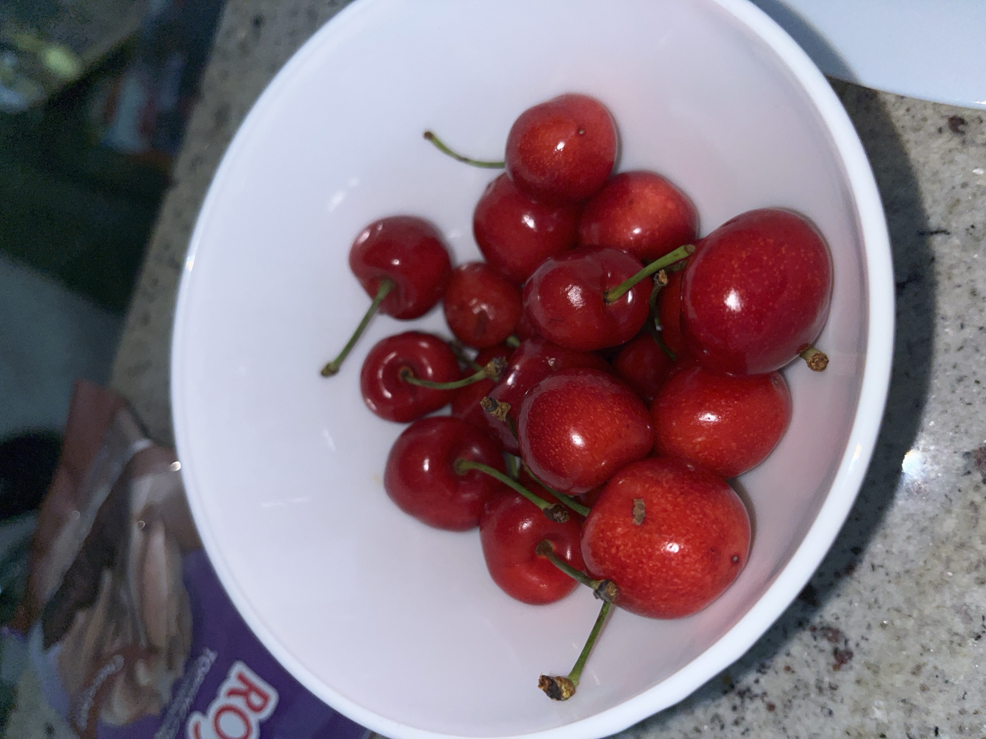 The image depicts an indoor scene in the city of Zlatograd, Bulgaria, focusing on a white, round ceramic bowl filled with approximately 20-30 ripe red cherries. The cherries vary in hue from bright crimson to darker maroon, with many retaining their light green or brown stems. Some cherries exhibit small, lighter speckles on their skin, and reflections of a light source are visible on their glossy surfaces. The bowl rests on a speckled countertop, which appears to be composed of granite or a similar material, displaying a mosaic of grey, black, and light brown flecks. In the blurred background to the left of the bowl, a portion of a purple package is visible, with the letters "ROY" clearly discernable, suggesting part of a brand name. Above this package, an indistinct, blurred object with light brown tones is present. The background to the right is dark and out of focus, with a faint, elongated light reflection on the far right edge. No individuals are present in the frame, and the cherries are static within the bowl.