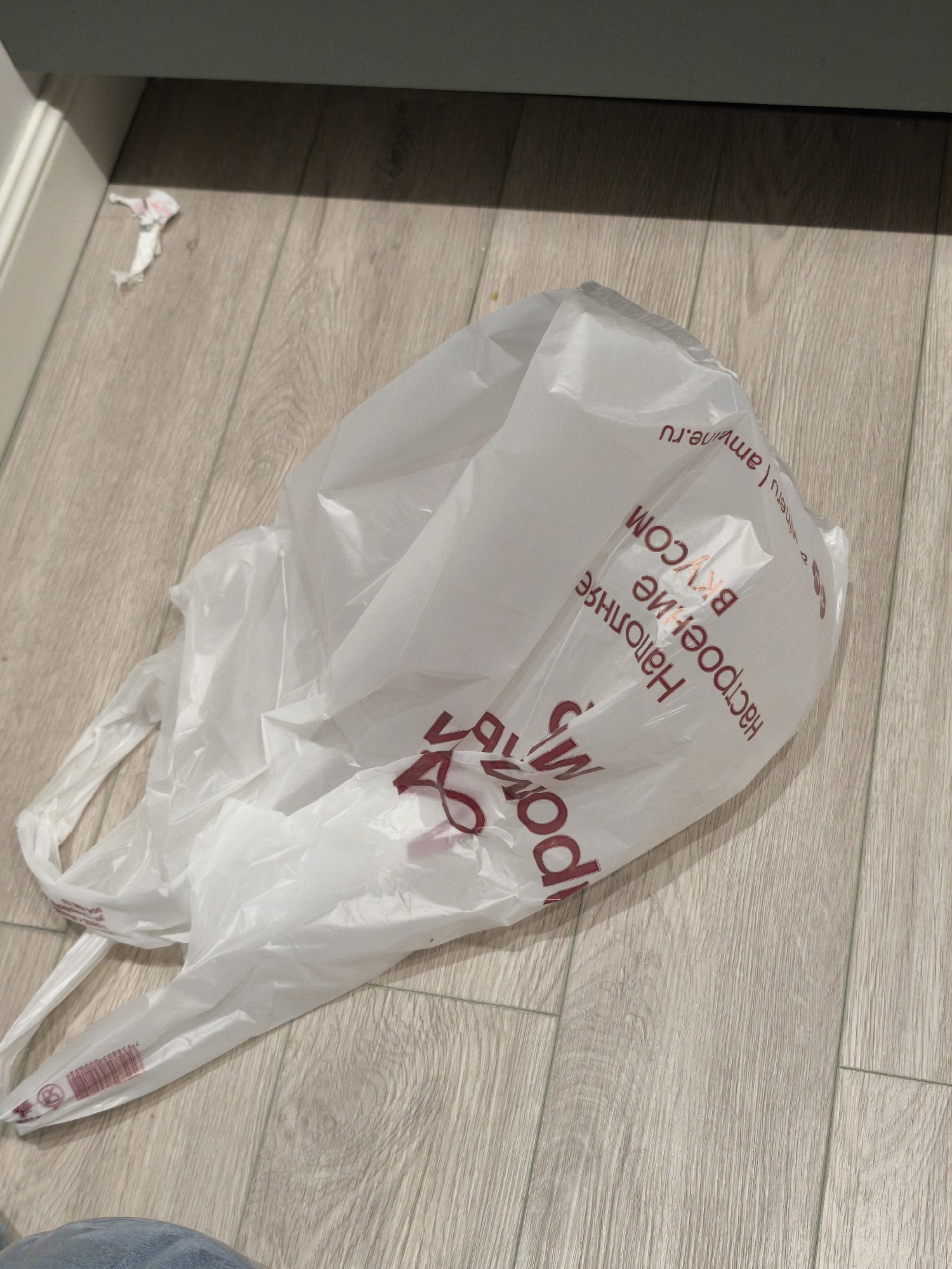 The image shows a white plastic shopping bag lying on a light-colored wooden floor. The bag has red text in Russian, including the words "РОССИЯ" (Russia) and "Наполнение вкусом" (Filling with taste). Part of a blue-colored garment is visible at the bottom left corner of the image. In the background, a piece of white tissue paper is visible, and the wall is painted a pale greenish-gray. Considering the text on the bag, the location is likely in Moscow, Russia.