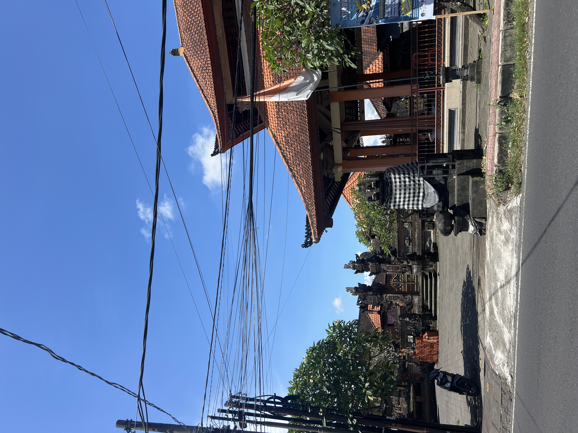 The image captures a street scene in Singapadu Tengah, Indonesia. It features traditional Balinese architecture, visible in the ornately decorated temples and buildings with red-tiled roofs. A power pole is visible in the foreground, with numerous electric wires stretching across the sky. A small motorcycle is parked along the street. The sky is clear and blue. A sign with some writing, and a piece of cloth hanging from one of the buildings. The perspective is angled slightly upwards, emphasizing the height of the structures. There are black and white checkered cloths draped over the statues.