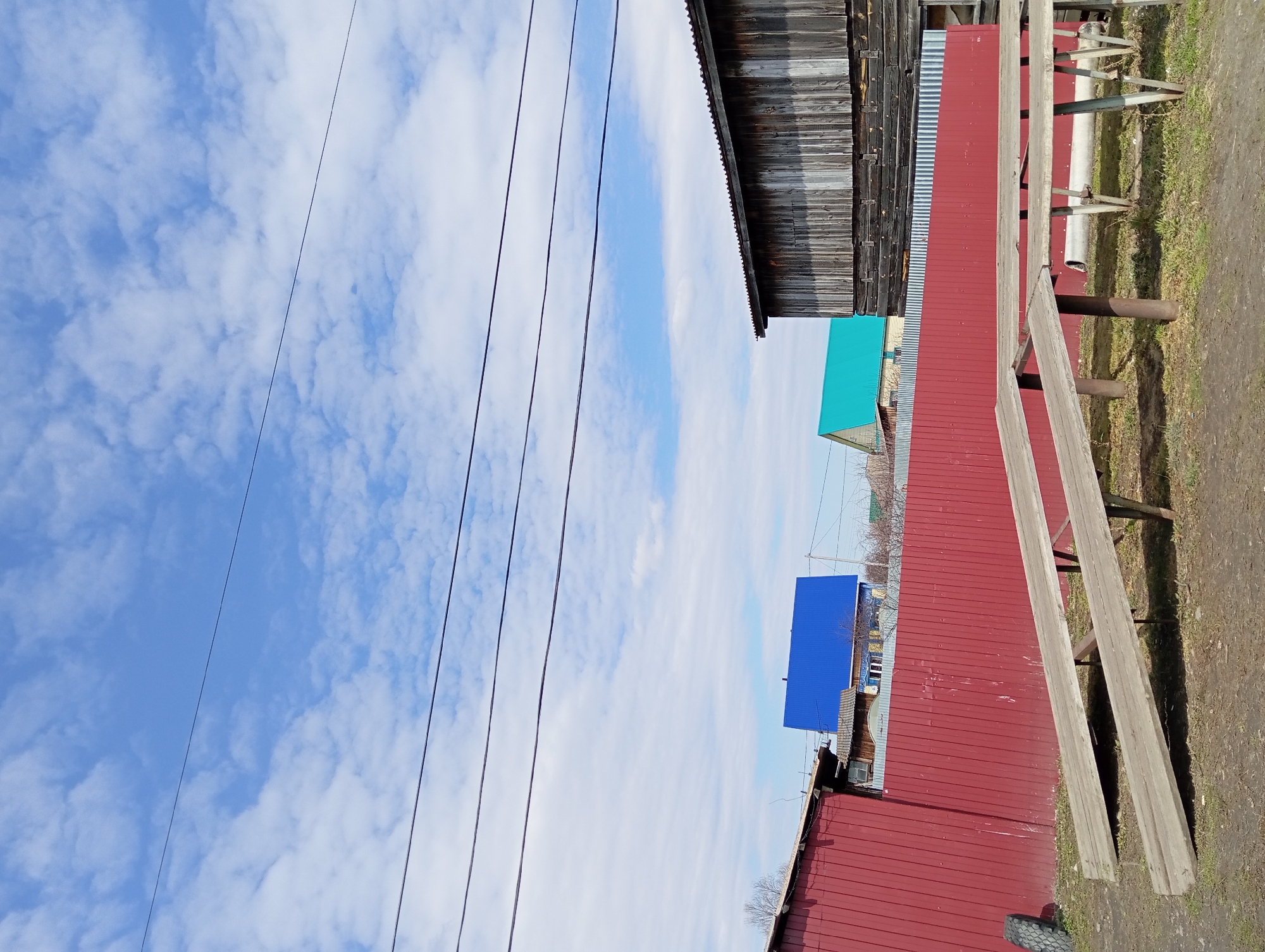 The image depicts an outdoor scene in Yurgamysh, Russia. Power lines stretch across a blue sky with scattered clouds. Adjacent to the sky, there is a variety of structures, including a dark wooden building with weathered siding, a red sheet metal building, and smaller buildings with blue and green roofs. A partially visible wooden bench is positioned near the sheet metal building. A dirt path borders the structures, with patches of grass visible.