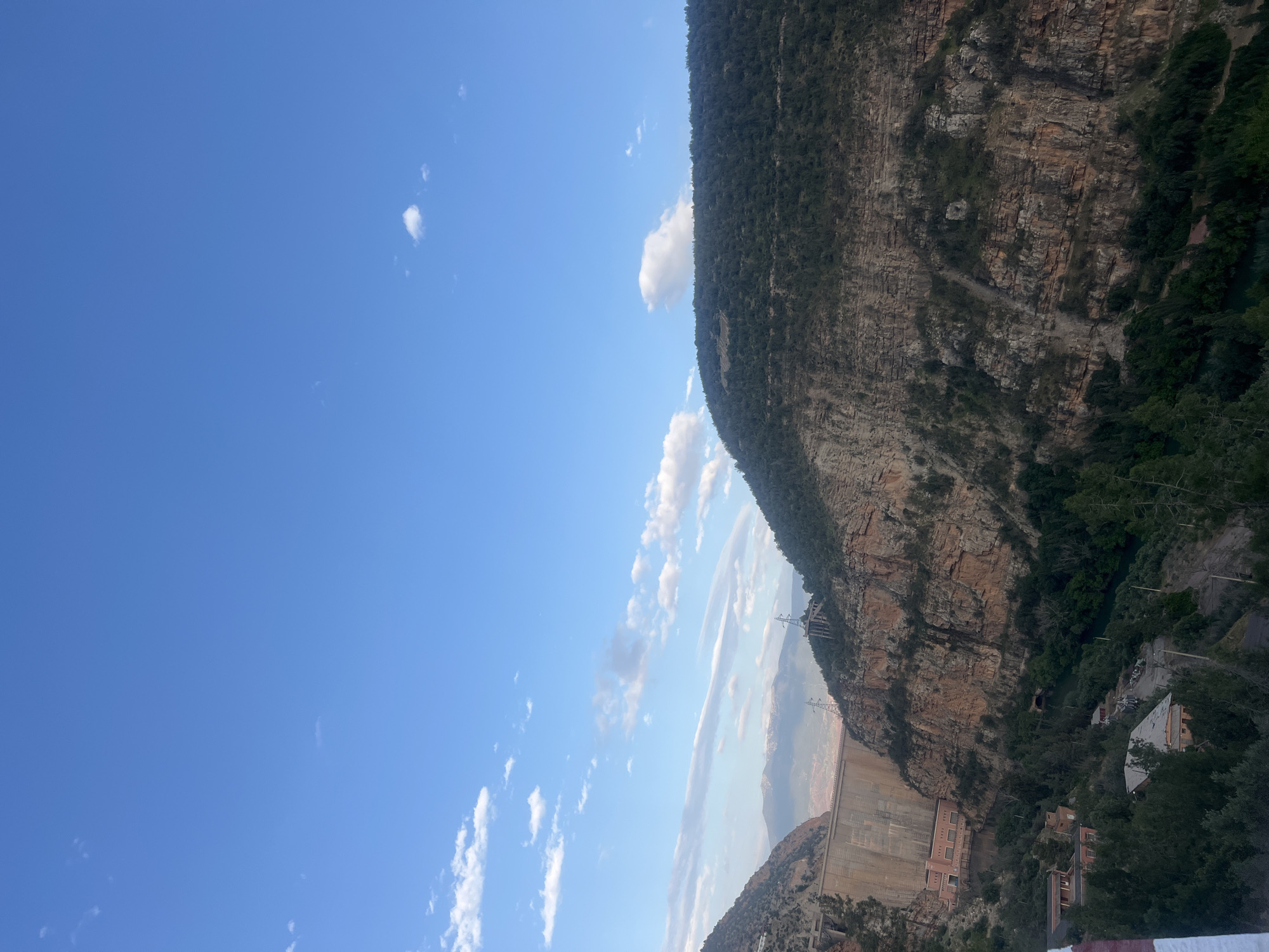 The image shows a partial view of Bin El Ouidane, Morocco, including the hydroelectric dam and a steep, rugged mountain rising behind it. The sky is blue with scattered clouds, and the view is from an elevated position. The cliff face of the mountain displays textures of rock and vegetation. Buildings and foliage are visible at the base of the dam and around the mountain. Electrical transmission towers are situated on the cliffside.