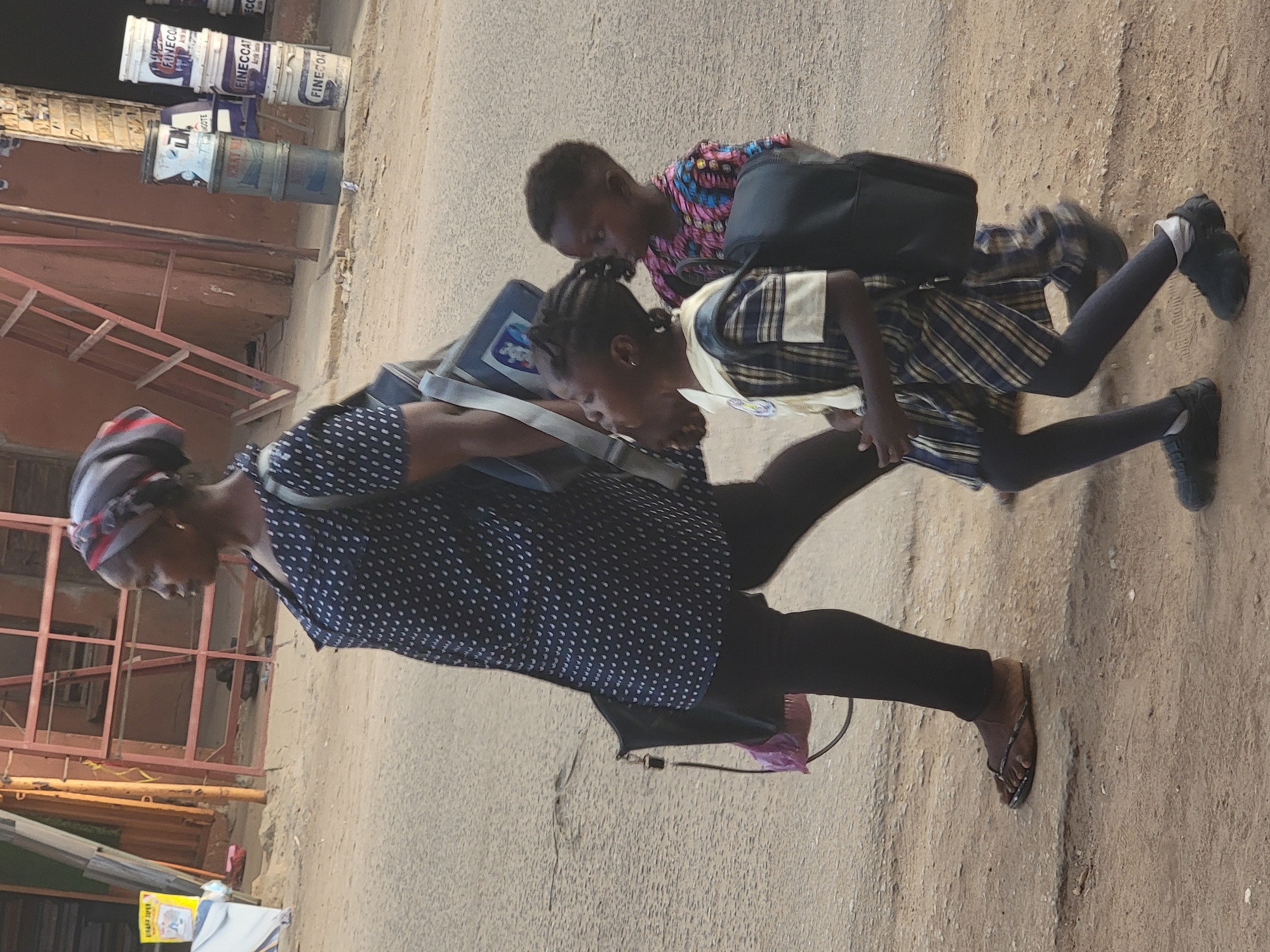 In Maiburiji, Nigeria, a woman in a polka-dot shirt, black leggings, and sandals walks on a paved street alongside two children. The children are wearing school uniforms and carrying backpacks. The surroundings consist of buildings, paint buckets stacked near a building, and metal fixtures along the wall. The scene appears to be a typical street with a mix of commercial and residential elements.