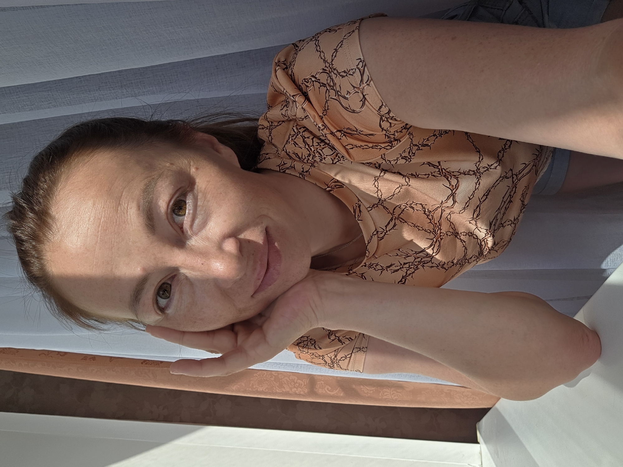 A Caucasian woman with medium brown hair, pulled back into a ponytail, is pictured in a selfie. She has a light complexion with freckles and brown eyes. She is resting her chin on her left hand, with her elbow on a white surface. The woman is wearing a light orange t-shirt with a black chain-like pattern. The background consists of beige and white curtains. The image appears to be taken indoors, possibly near a window, given the lighting and shadows cast. The location is Chaikovsky, Russia.