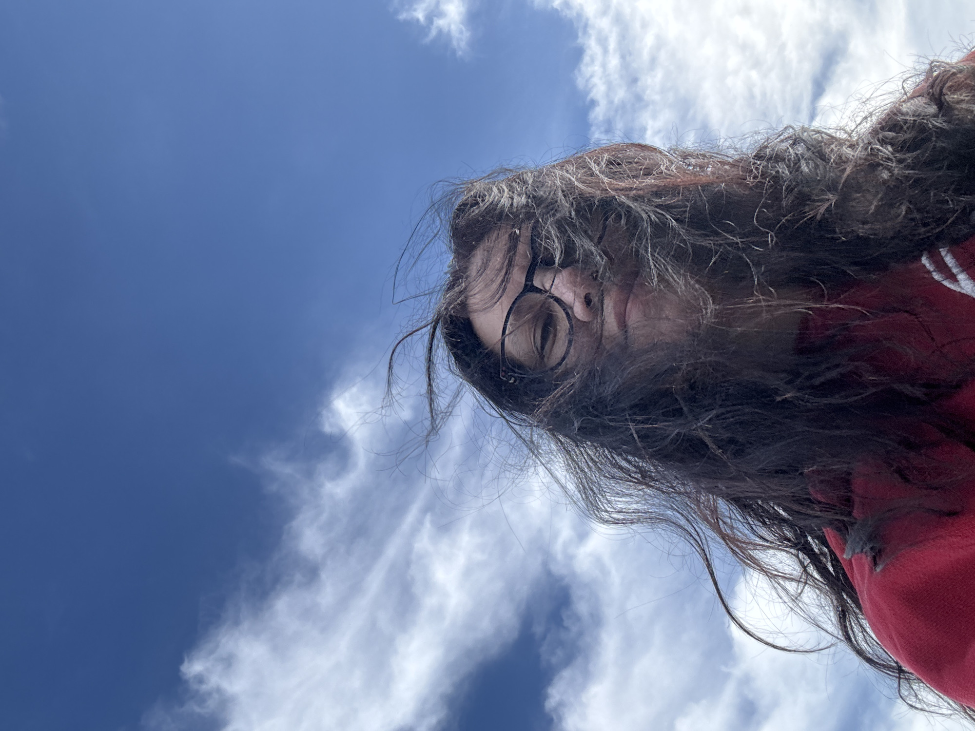 The image shows a low-angle shot of a person with long, dark hair and glasses, against a blue sky with scattered white clouds. The person is wearing a red garment. The location is not discernible from the image. There is no indication that this is Chaikovsky, Russia.
