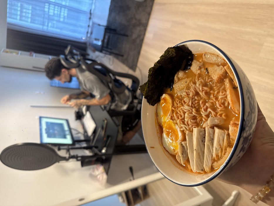 The image displays a hand in the foreground holding a white bowl with a blue patterned rim, filled with ramen. The ramen consists of orange broth, instant noodles, two halved soft-boiled eggs with visible orange yolks, multiple slices of white meat with black flecks, and a piece of dark green nori (seaweed) standing upright. A beaded bracelet with yellow-orange translucent beads is visible on the wrist holding the bowl.
In the mid-ground and background, an apartment interior in Bangkok, Thailand, is visible. A male individual with dark hair and a beard, wearing a grey t-shirt and shorts, is seated in a black ergonomic office chair at a black desk. He is looking towards the right, appearing to be manipulating an object with his hands. On the desk, a computer monitor displays a graphical user interface resembling a digital audio workstation, alongside a white keyboard, a mouse, and audio recording equipment including a pop filter and microphone stand. The apartment features light wooden flooring, white walls, and a large window with sheer white curtains on the right side, through which an adjacent urban high-rise building is visible. A dark grey rug and a small, dark rectangular table are present on the floor in the background.