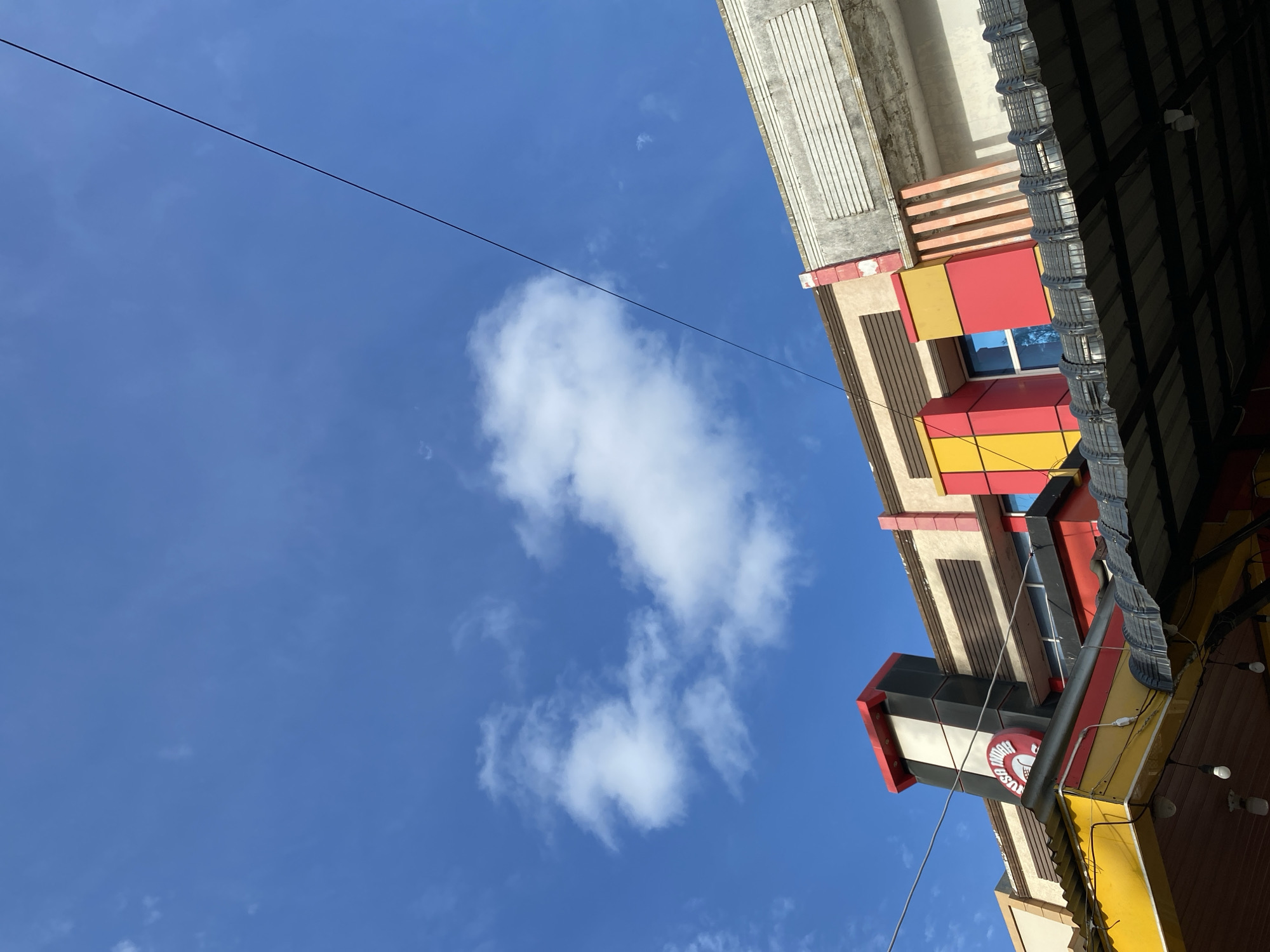 The image shows a low-angle view of a building's facade against a bright blue sky with scattered white clouds. The building features a vibrant design with sections of red, yellow, and black, contrasting with neutral beige and grey elements. A prominent circular sign with red lettering is visible on a black and red pillar at the top left of the building. Below this, a corrugated metal roof extends outwards, supported by beams and partially obscuring the lower part of the building. Electrical wires are strung across the sky, one of them running diagonally from the top right towards the center of the frame. The architectural style suggests it could be a commercial establishment. The location is identified as Lampisang, Indonesia.