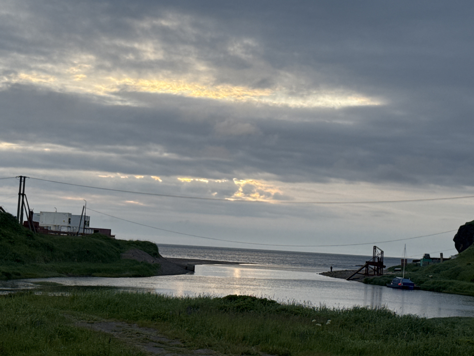 The image shows a body of water, likely a bay or inlet, with a gradual transition to the ocean. The sky is overcast with dramatic clouds, some of which are illuminated by sunlight breaking through, creating streaks of yellow and white. The water reflects the sky, adding to the muted, atmospheric quality of the scene.
Along the left bank of the water, there is a grassy hill with a few buildings. The buildings appear to be industrial or storage structures, painted white and red, with some utility poles and wires visible. The ground is covered in lush green grass and a dirt path is visible in the foreground.
On the right bank, the land rises steeply to form a rocky cliff. A small boat with a blue canopy is moored in the water near the shore. There is a wooden structure, possibly a dock or pier, with a ramp leading down to the water. Further back, more small buildings and utility poles are scattered along the hillside. A solitary figure stands on the shore near the wooden structure.
The overall impression is a remote, coastal settlement with a functional, perhaps industrial, character. The lighting suggests either early morning or late evening. The scene is evocative of a northern, rugged landscape, consistent with the location in Kuril'sk, Russia, known for its volcanic terrain and maritime environment.