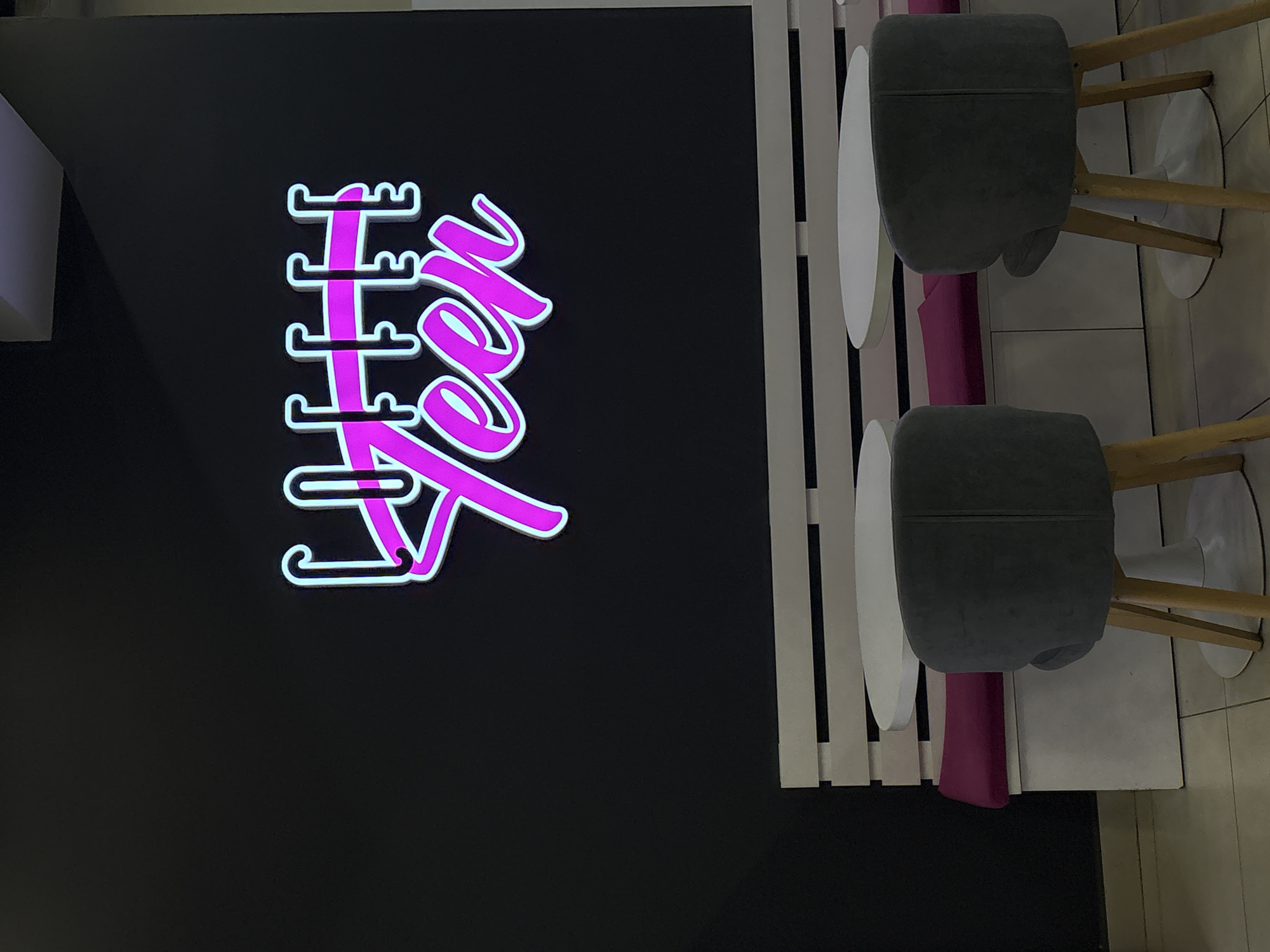 The image depicts a cafe interior. A neon sign on a black wall reads "COFFEE TEEN" in bright pink and blue. Below the sign is a white bench with a fuchsia cushion. Two small white tables are positioned in front of the bench. Two grey upholstered chairs with wooden legs face the tables. The floor is tiled. The scene appears to be an interior shot of a cafe.