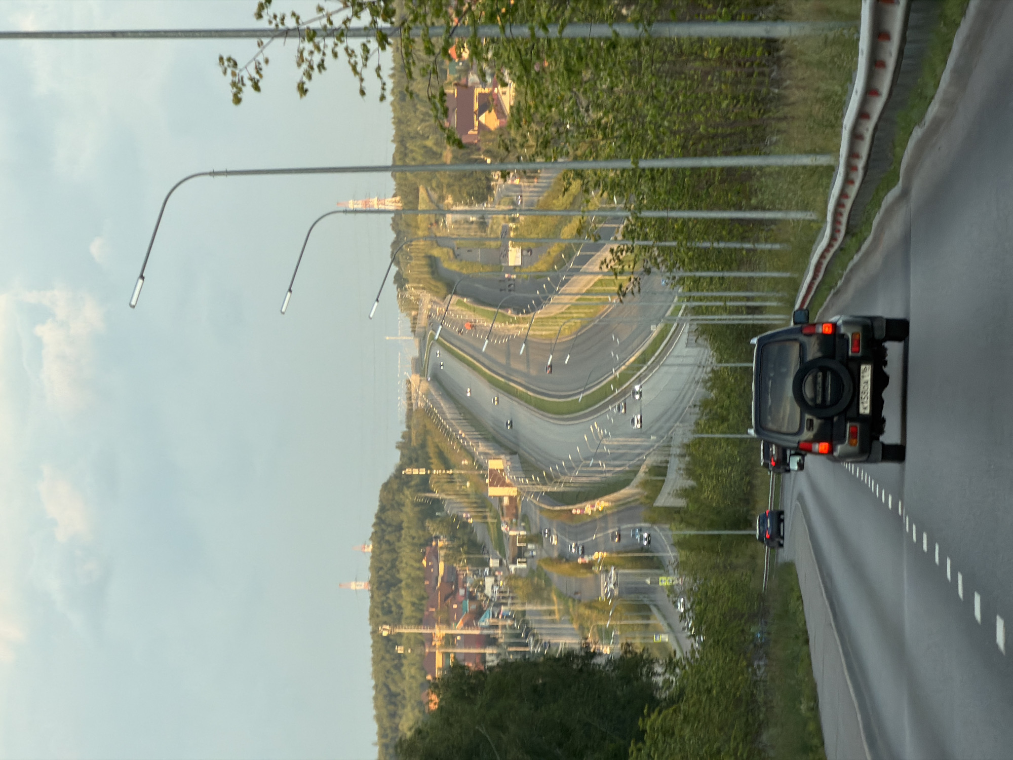 This image captures a view of a modern, multi-lane highway curving through a suburban landscape on the outskirts of Kazan, Russia. The highway is lined with streetlights, and several vehicles are visible on its lanes. In the foreground, a dark-colored SUV is driving away from the camera. Beyond the highway, residential buildings with red-tiled roofs are nestled among lush green trees covering rolling hills. In the distance, two tall industrial towers with red accents are visible on a hilltop, and a bridge can be seen in the far background. The sky above is partly cloudy, with soft, diffused light suggesting either early morning or late afternoon. The overall impression is one of a developing urban area integrated with a natural, hilly environment.