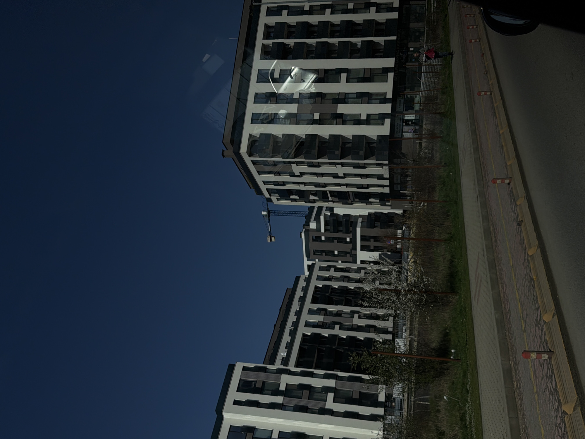 The image shows a modern apartment building in Sofia, Bulgaria, viewed from a moving vehicle. The building has a white facade with dark-framed windows and balconies. A construction crane is visible near the middle building. A sidewalk runs along the base of the building, and a street is to the right. A pedestrian is seen near the building's entrance. The sky is clear and dark blue.