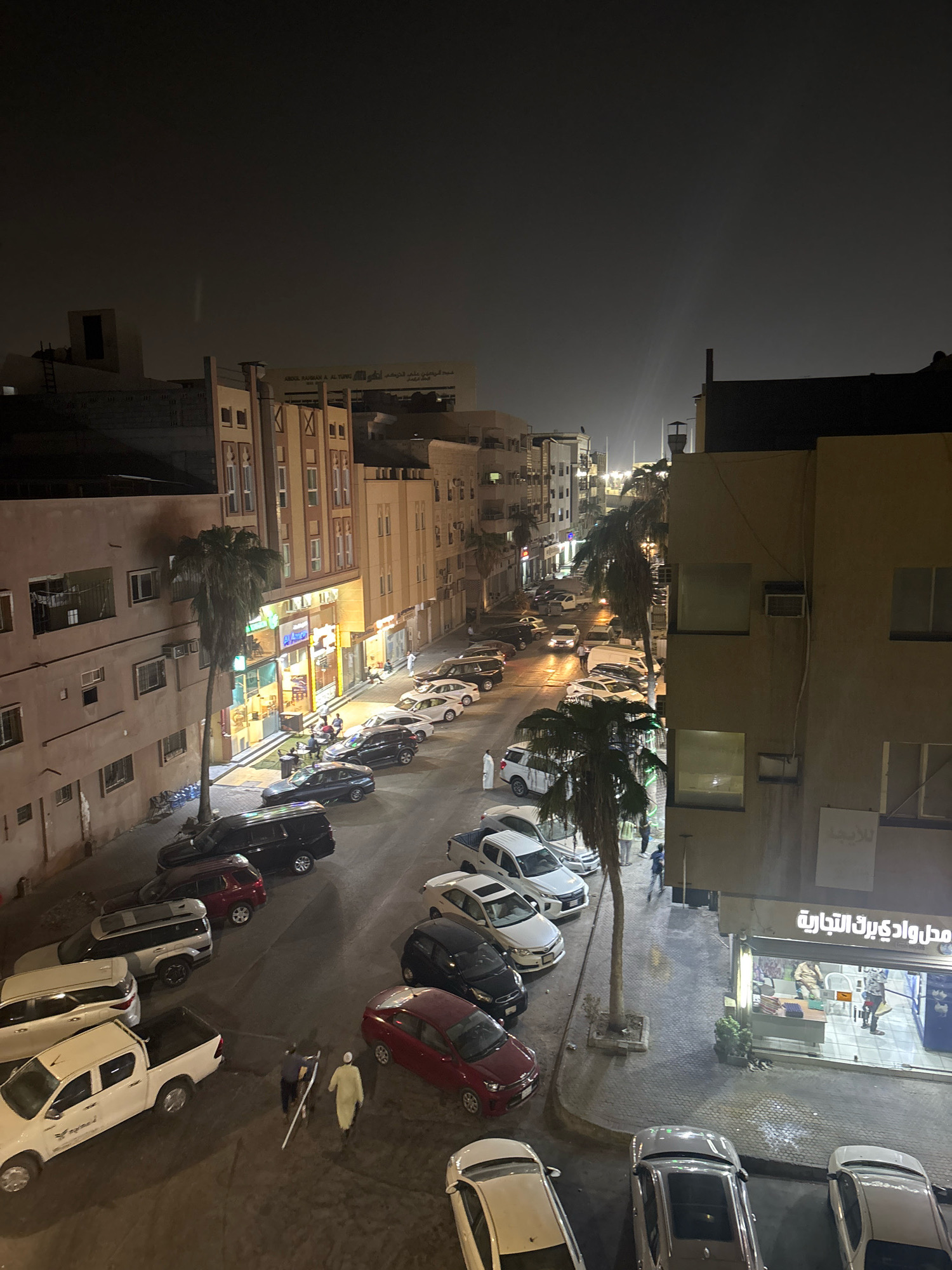 This is a high-angle, nighttime photograph of a street scene in Dammam, Saudi Arabia. The street is lined with multi-story buildings, some of which appear to be businesses with illuminated storefronts. Several palm trees are scattered along the street and sidewalk. A number of cars are parked on both sides of the street and on the sidewalk. There are a few people visible on the street, some walking and some gathered outside a storefront. The overall atmosphere suggests a busy urban environment at night.