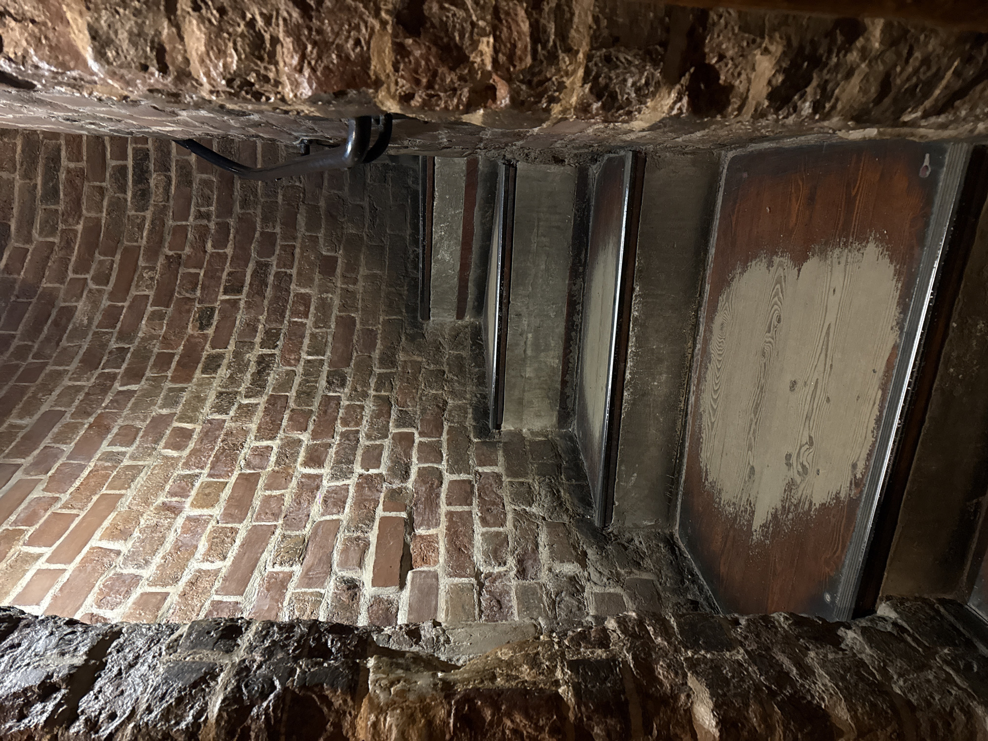 This image depicts a narrow, enclosed space, possibly a stairwell or passage, within a building. The space is constructed of a combination of materials: brick, wood, and metal. The walls are primarily brick, laid in a pattern. The ceiling and floor appear to be made of rough-hewn stone or brick. Steps, possibly made of wood and reinforced with metal edges, are visible. One of the wall sections is wooden. The lighting is dim, with shadows obscuring some details. There is a metal pipe running along the ceiling. There are no people in the scene. The location is Moscow, Russia.