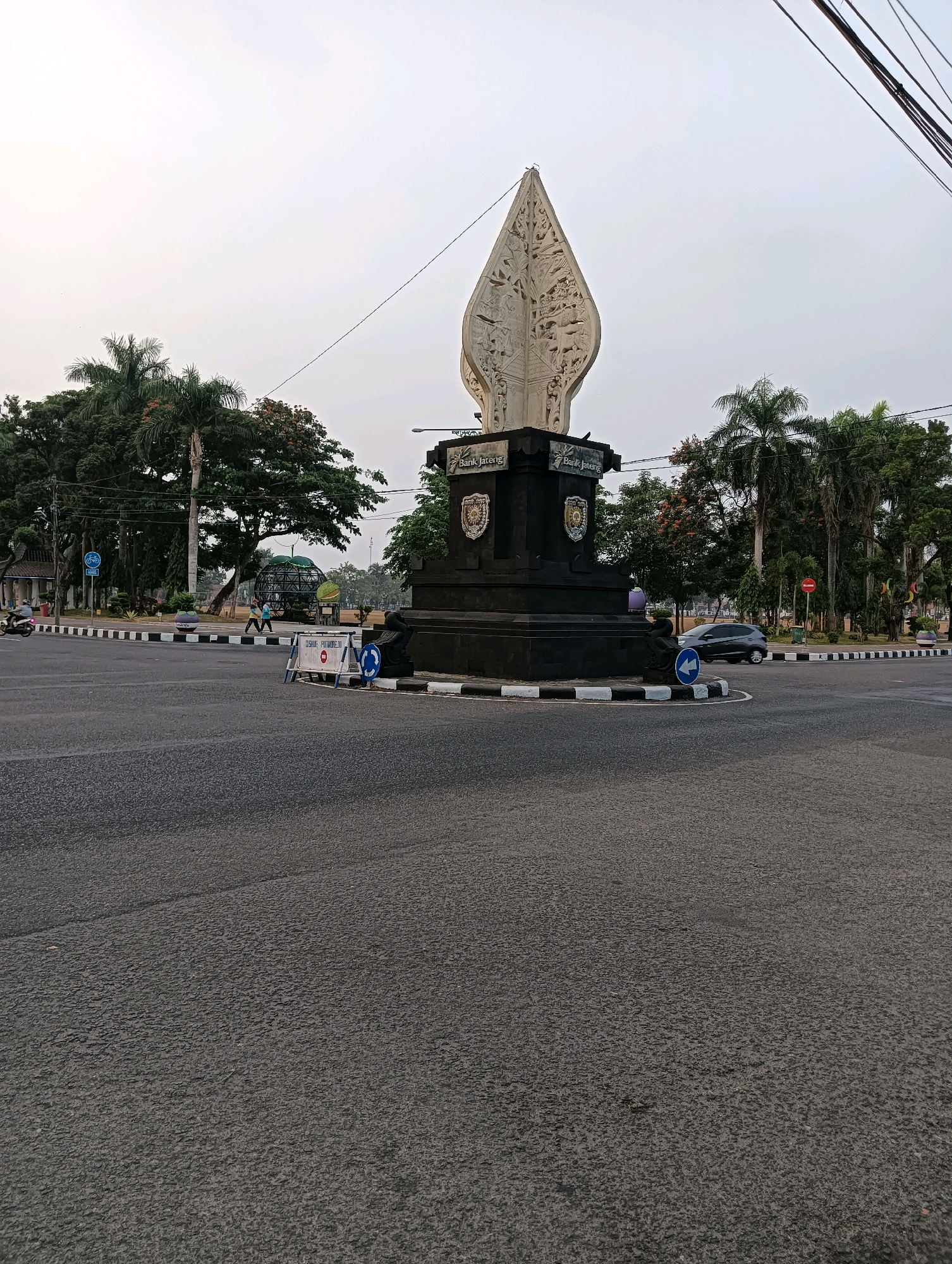 The image depicts the Tugu Pancasila (Pancasila Monument) in Purworejo, Indonesia, viewed from a low angle at an intersection. The monument, a prominent landmark, is tall and features intricate carvings at its apex. It is situated on a dark, multi-tiered base with inscriptions and emblems. Surrounding the monument are lush green trees, including palm trees, and a clear, overcast sky. The foreground is dominated by an asphalt road, marked with white and black stripes at the intersection. Traffic signs, including a blue directional arrow sign and temporary barriers, are visible on the road. Several cars and a motorcycle are present, indicating ongoing traffic. Two individuals are walking in the background, and one person is seated on the monument's base. The overall scene suggests a typical urban environment in Purworejo, likely captured during the daytime.