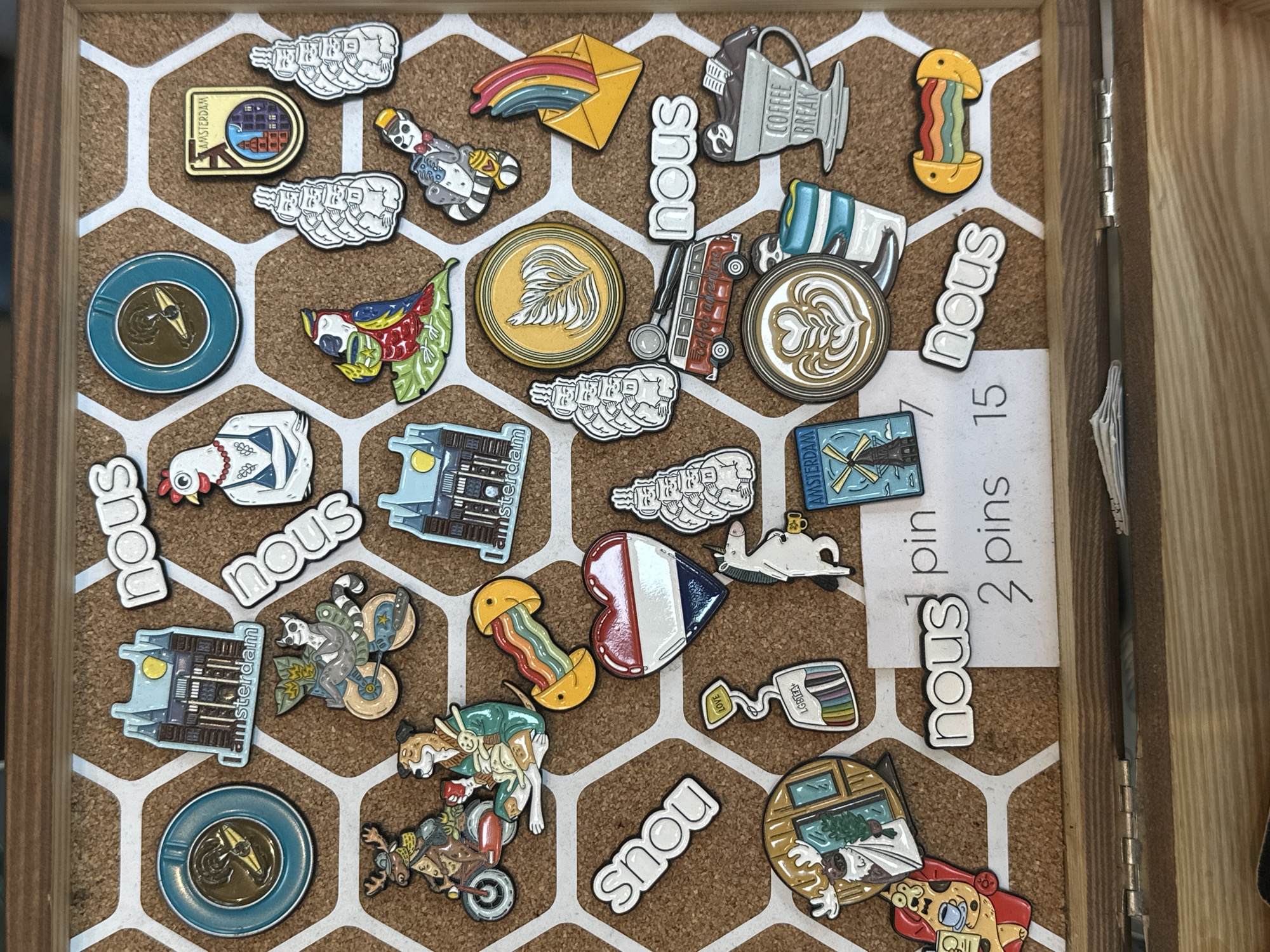 The image shows a close-up, slightly overhead view of a corkboard display, likely in a retail setting. The corkboard has a white hexagonal pattern superimposed on it. The board is covered with various enamel pins, many of which are themed around coffee, Amsterdam, and whimsical characters. Several pins depict iconic Amsterdam architecture, such as canal houses and a windmill, with one pin explicitly labeled "Amsterdam". Other pins feature animals like a moose, a dog, a raccoon, and a parrot, often engaged in human-like activities such as riding bicycles or drinking coffee. There are also abstract designs, such as a rainbow coming out of a yellow envelope and a pin with the word "nous" repeated. A small sign with handwritten text "1 pin 2 pins 3 pins 15" is also present. The display is contained within a wooden frame, with visible hinges at the bottom, suggesting it may be a lid or part of a larger display case. The lighting is even and appears to be from above, casting minimal shadows.