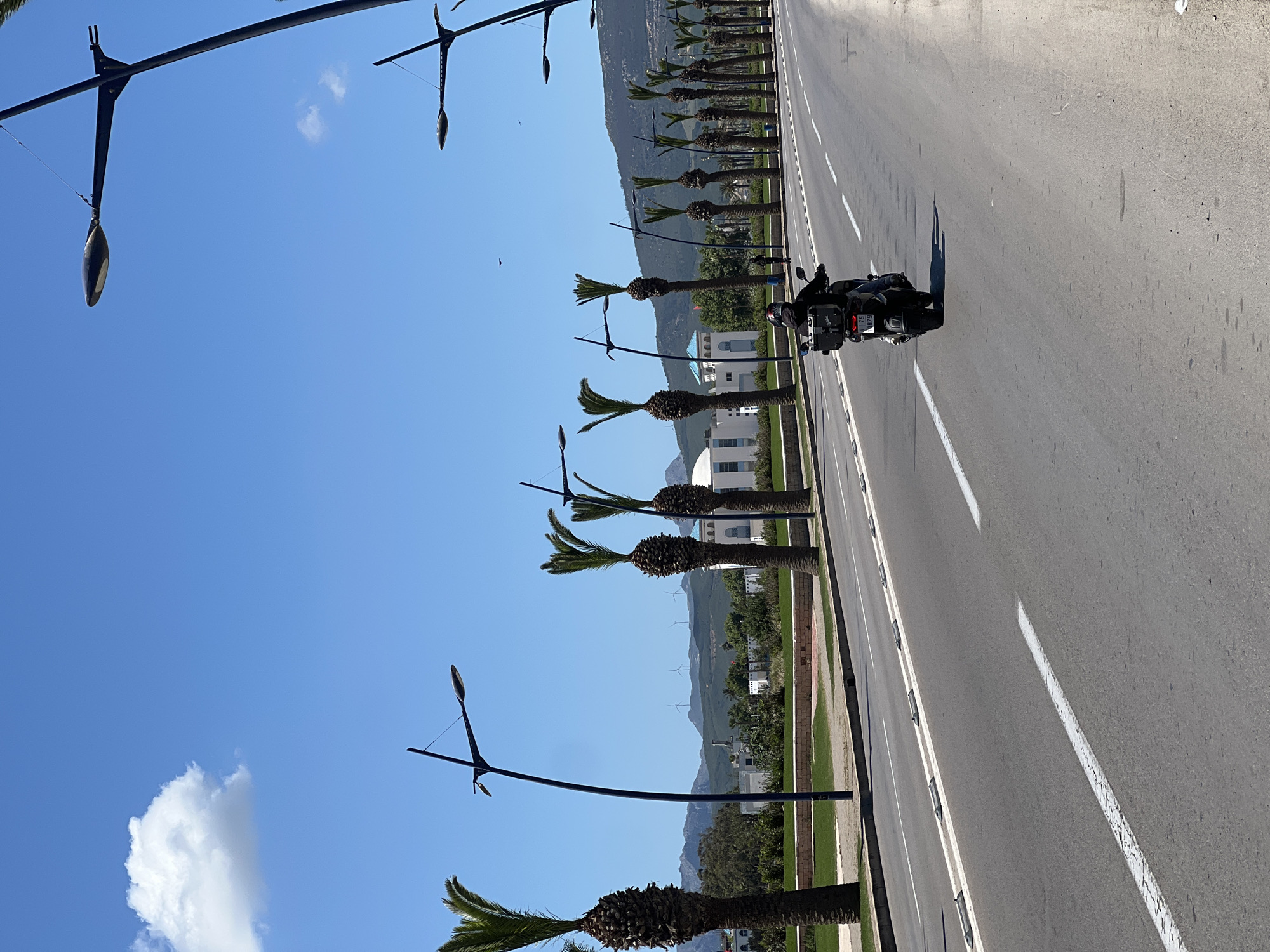 The image shows a motorcycle moving on a road lined with palm trees and streetlights. In the background, there are buildings and mountains. The sky is blue with some clouds. The location is M'Diq, Morocco.