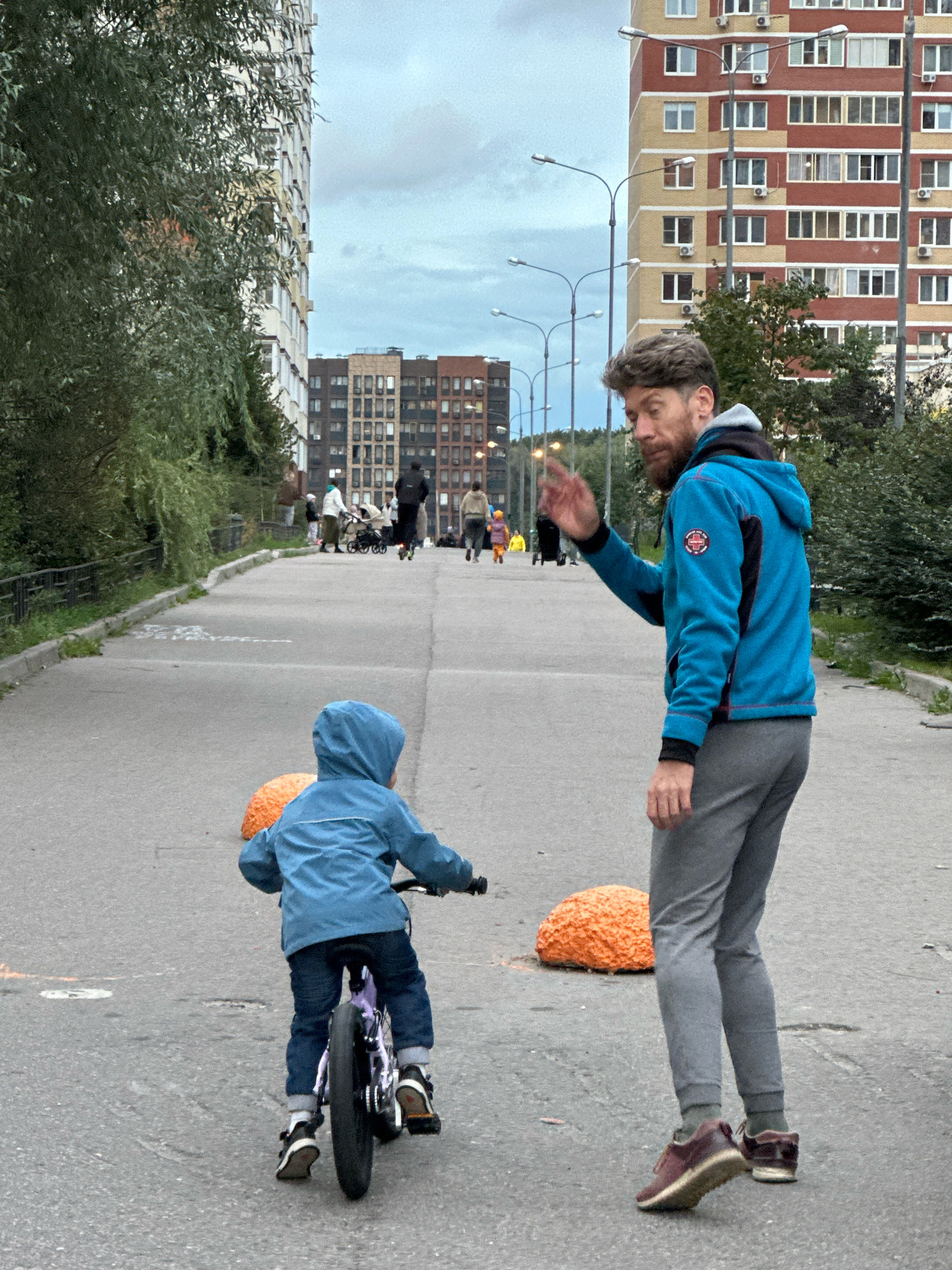 A man and a child are on a paved road in an urban environment. The man, with a beard and short hair, wears a blue hooded jacket and gray pants. He is facing away from the camera, looking towards the child, and has his right hand raised as if waving or gesturing. The child, wearing a blue hooded jacket and dark pants, is riding a small bicycle in front of the man, also facing away from the camera. Two large, orange, textured objects are visible on the road near the child. In the background, tall apartment buildings line the road, and several streetlights are present. Other people and children are visible in the distance. The scene appears to be in Vidnoye, Russia.