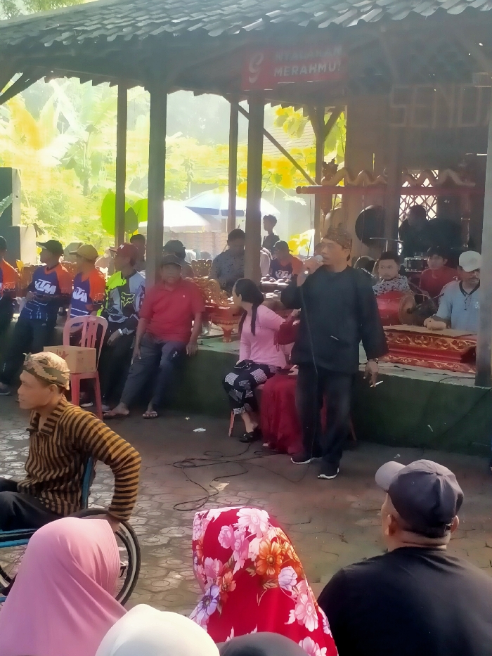 A group of people are gathered under a roofed structure in Masaran, Indonesia (latitude: -7.471, longitude: 110.957). A man in a black shirt and a black cap is speaking into a microphone. He is standing in front of a red-and-gold traditional music instrument. Behind him, seated on a wooden platform, are several more people. Among them are a woman in a pink dress and a man in a white shirt with a black collar. In the foreground, a man in a striped shirt is sitting in a wheelchair. Other people, including a woman in a pink headscarf and a woman in a red headscarf, are seated in the front row. Green foliage can be seen in the background.