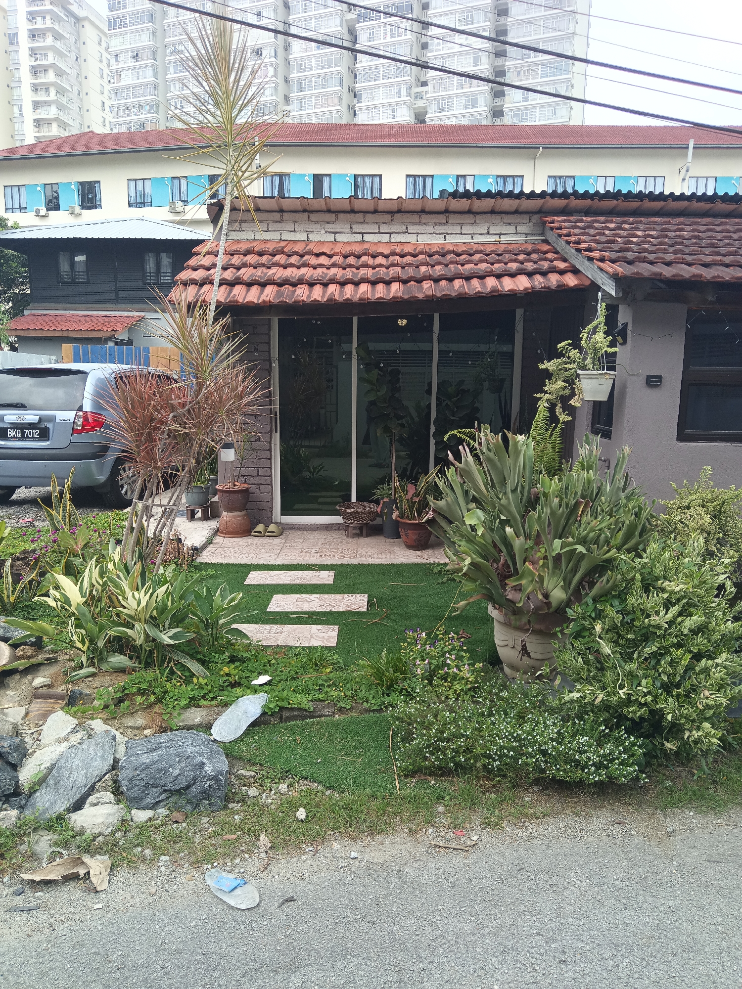 The image shows a residential property in Kuala Lumpur, Malaysia, on an overcast day. In the foreground, a grey car with license plate BKQ 7012 is parked to the left. A path of light-colored stepping stones is laid across a patch of artificial turf, leading to the entrance of a single-story house. The house has a terracotta-tiled roof and a modern facade with large glass sliding doors. The entrance is adorned with various potted plants, including a large staghorn fern in a decorative pot. To the left of the house, a garden area features rocks, small plants, and a Dracaena plant. In the background, a multi-story apartment building with numerous windows dominates the skyline. Overhead power lines are visible. The general environment suggests a residential area with both older and newer constructions.