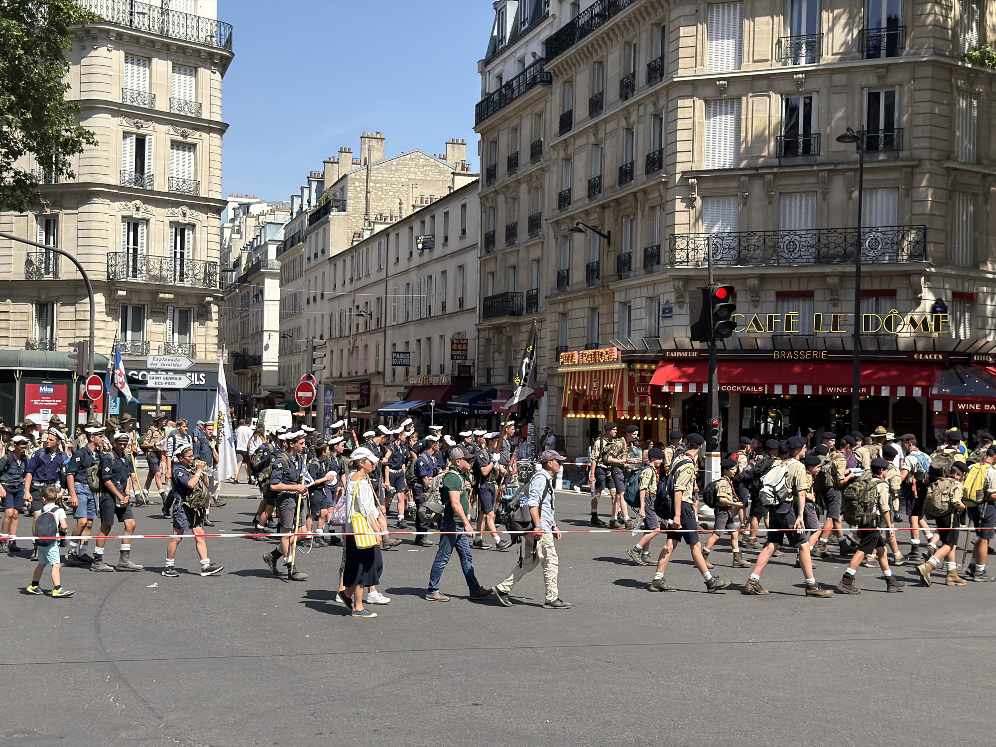The image shows a large group of people, primarily children and young adults, marching in a procession across a street in Paris, France. Many of the people are wearing uniforms that suggest they are members of a scouting organization. They are carrying backpacks and flags. The procession is crossing a street that is marked off by tape. Behind the procession are buildings typical of Parisian architecture, with multiple floors and balconies. The street is lined with commercial establishments including the "Cafe Le Dome" brasserie. Street signs and other commercial signage are visible. The street signs include "Esplanade des Invalides SAINT GERMAIN DES PRES", "PARIS SOUVENIRS SAVEURS D'ASIE", and "PATISSERIE". A red traffic light can be seen near the Cafe Le Dome. The weather appears to be sunny and bright.