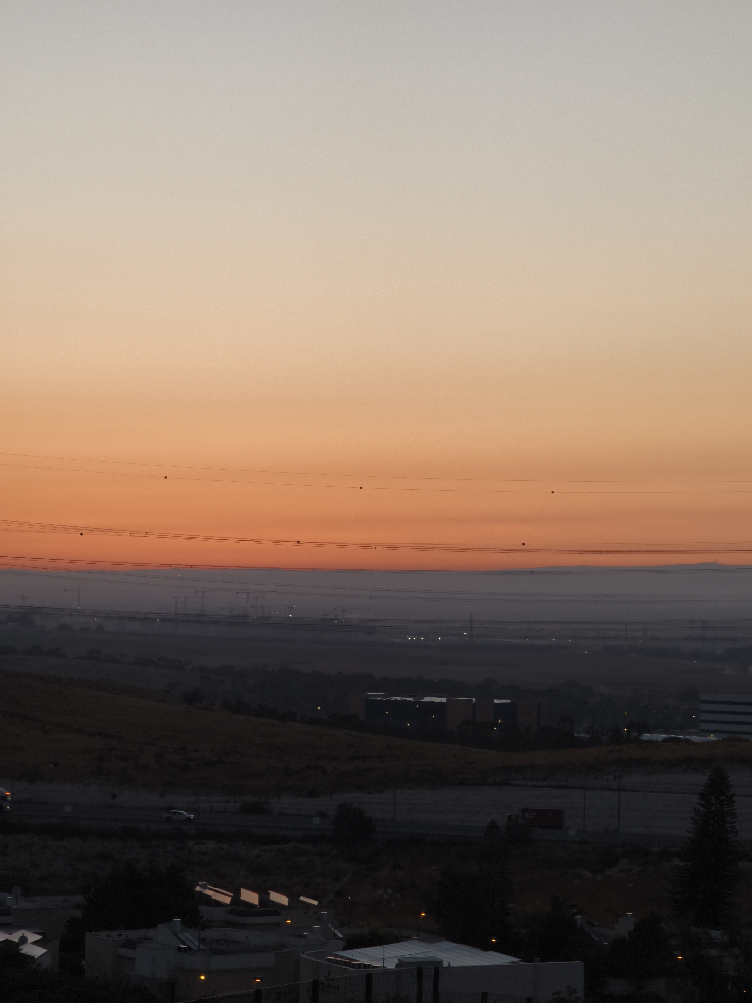 This image depicts a wide, horizontal landscape scene at dusk, featuring a gradient sky transitioning from light grey at the top to a vibrant orange near the horizon. A series of power lines crisscross the sky, with small, dark circular insulators visible on them. Below the sky, a hazy band of lighter grey stretches across the midground, obscuring the distant landscape. In the foreground, the scene transitions to a darker, silhouetted environment. On the left, a hilly, dry, golden-brown landscape is visible, leading to a residential area with low-rise buildings. Several of these buildings have solar panels installed on their roofs, reflecting the dim light. A white SUV is driving on a road in the mid-left of the frame, and a red truck is visible further to the right on a road in the darker foreground. The overall impression is a panoramic view of the outskirts of Be'er-Sheva, Israel, as daylight fades.