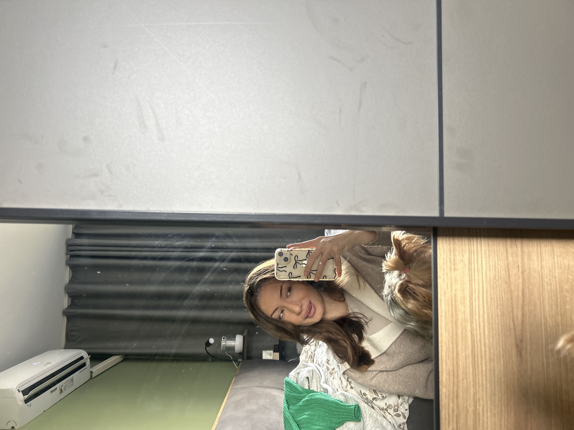 The image shows a reflection in a mirror. The reflection depicts a young woman holding a smartphone in her right hand, likely taking a selfie. She has brown hair and is wearing a light-colored top and a cardigan. Behind her in the reflection is a room with gray curtains, a green surface, and an air conditioning unit. There's also what appears to be a bed or couch with some clothing on it. A small dog is near the woman's head. The location is unknown and cannot be determined from the image.