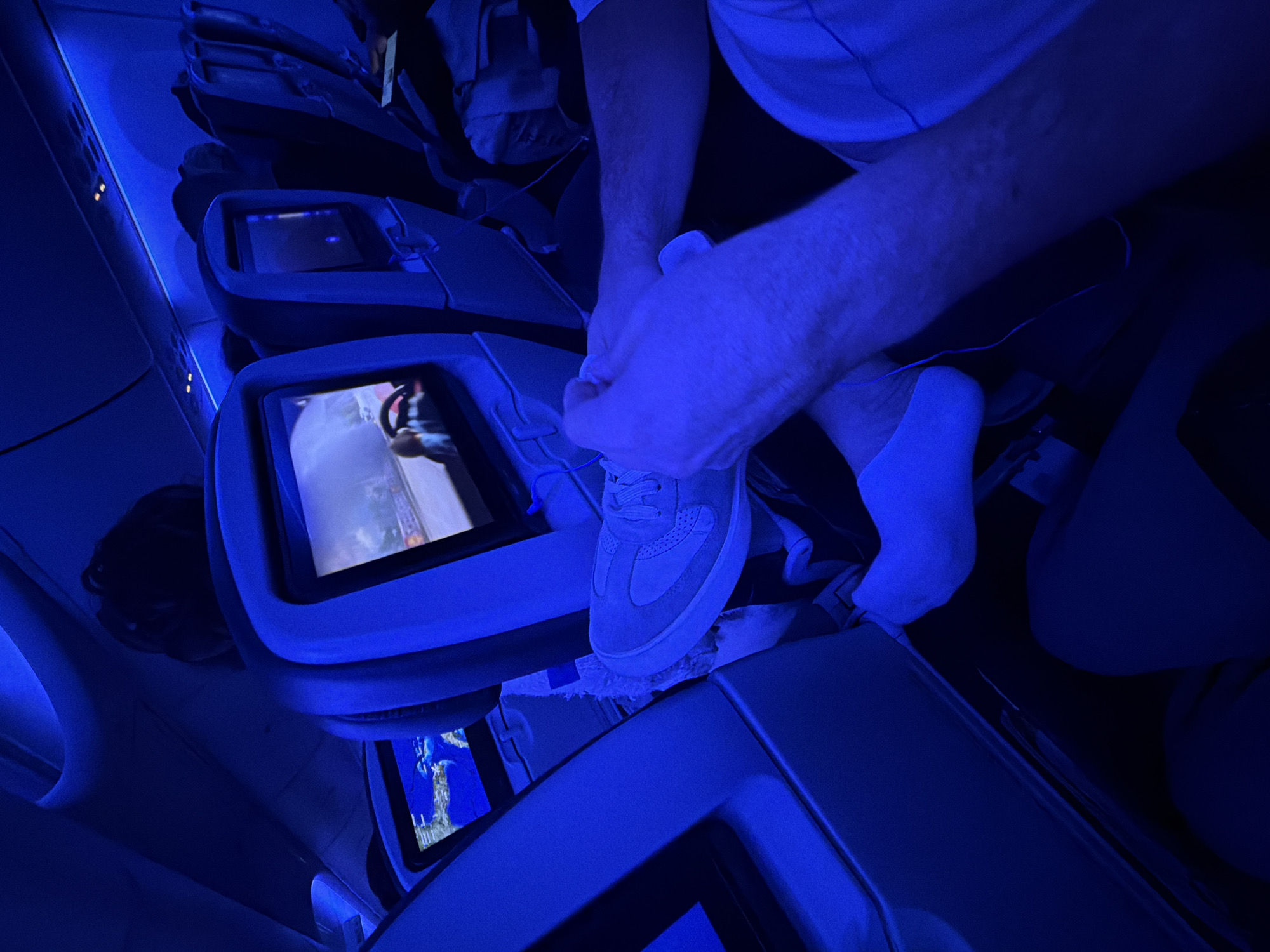 A person is shown tying a shoe in the seating area of an airplane. The airplane's interior lighting is predominantly blue. The person is wearing white socks and is tying the laces of a light-colored sneaker. The airplane's entertainment system is visible on the seat in front. The location in Miami, United States of America, cannot be determined from the provided visual data.