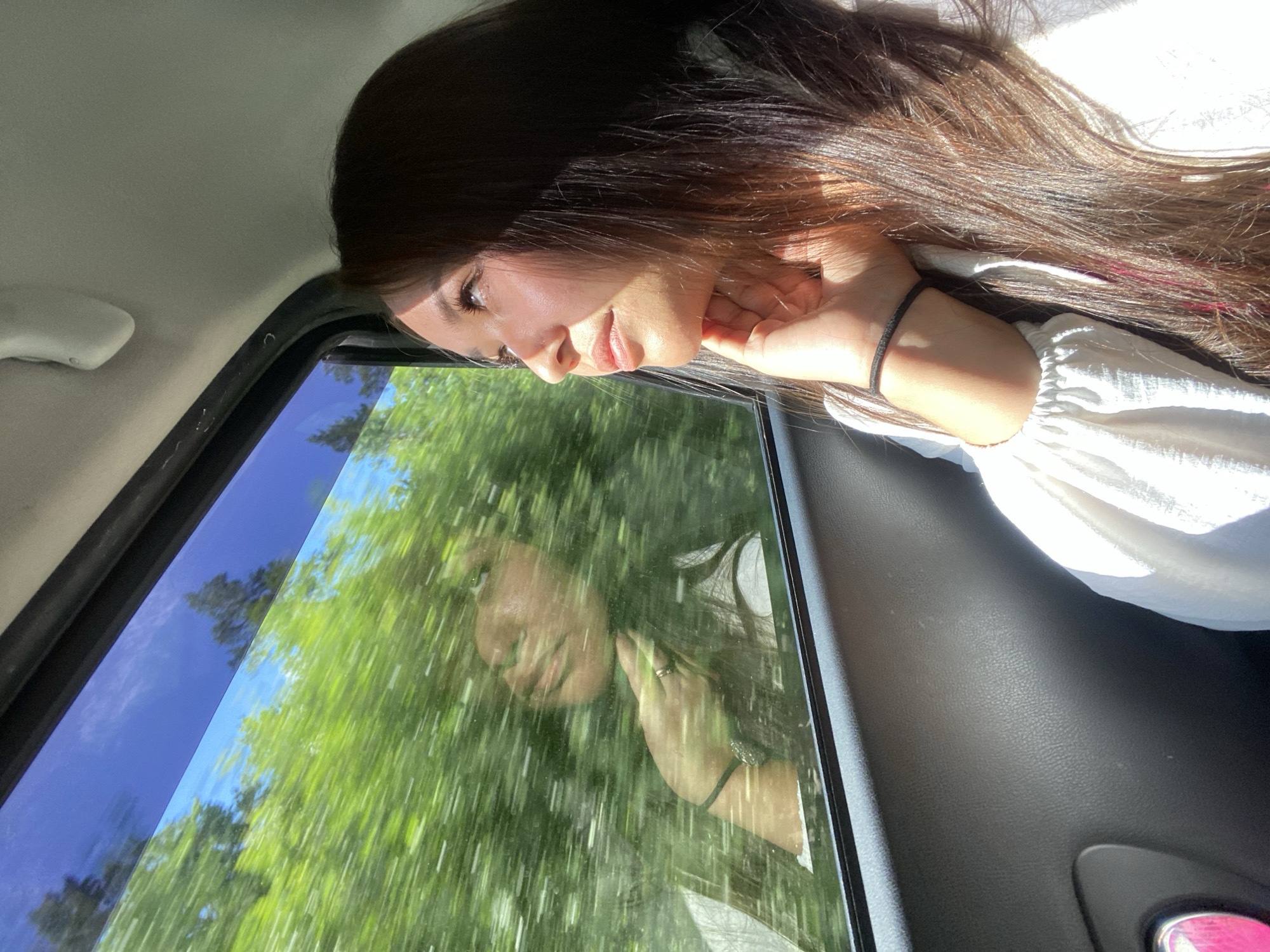 A young woman with long, dark hair and fair skin looks out a car window, her hand resting on her chin. Her reflection is visible in the window, superimposed over a blur of green trees and blue sky, suggesting the car is in motion. She is wearing a white blouse. The interior of the car is visible, including the door panel and a small pink object. The scene takes place during daylight. The question about Zilair, Russia cannot be answered as there is no information in the image to suggest this location.