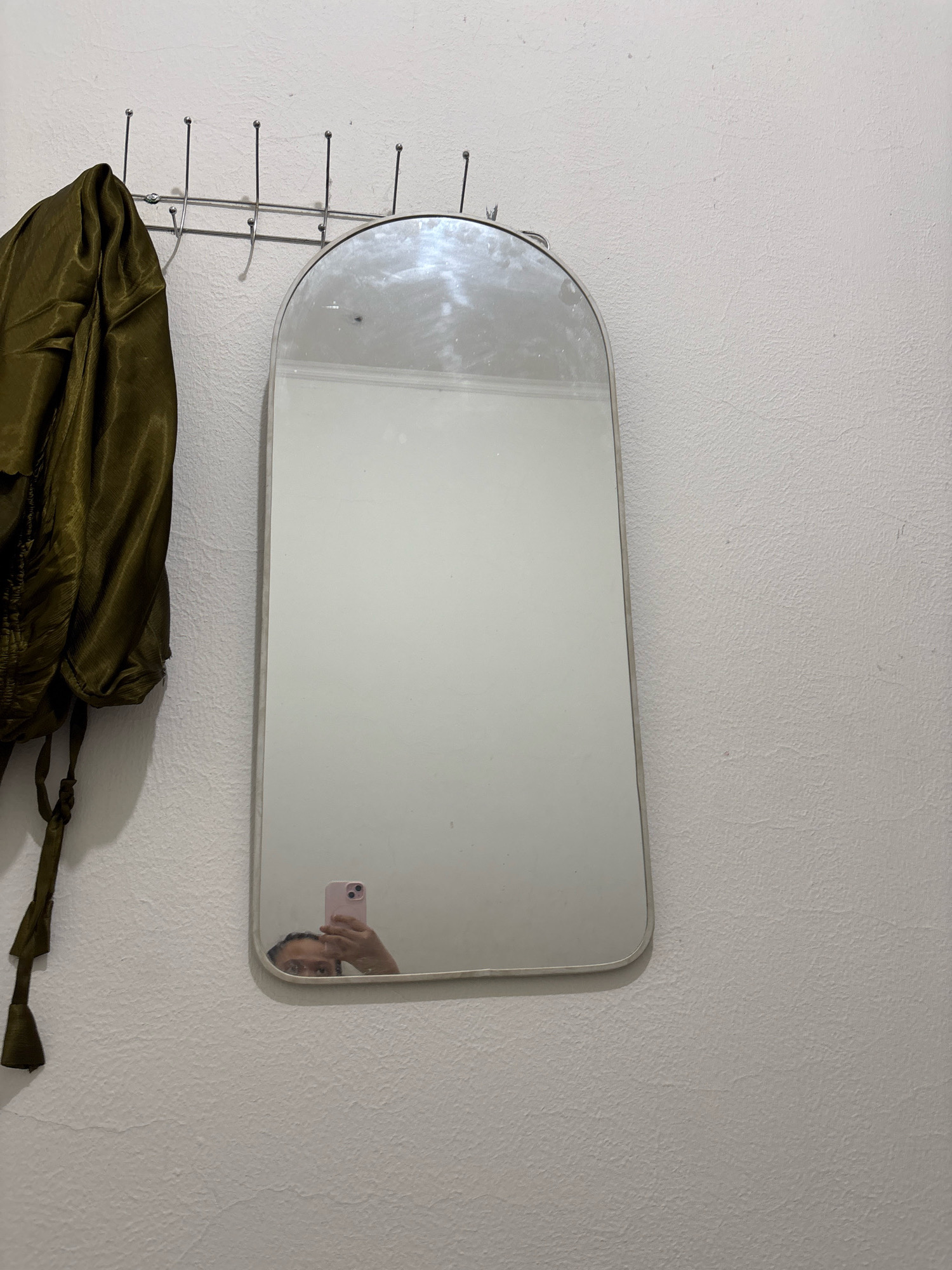 A mirror with a rounded top is mounted on a textured white wall. To the left of the mirror, a dark olive green jacket hangs from a metal coat rack with multiple hooks. The jacket appears to be made of a shiny, rumpled fabric. In the reflection of the mirror, a person is visible holding a pink smartphone up to take a selfie. The person's face and upper body are reflected, along with the phone screen. The background of the mirror reflection is the interior of a room, but it is out of focus and appears bright. The scene is indoors, likely in a residential setting, and based on the location, it is in the city of Wanajaya, Indonesia.