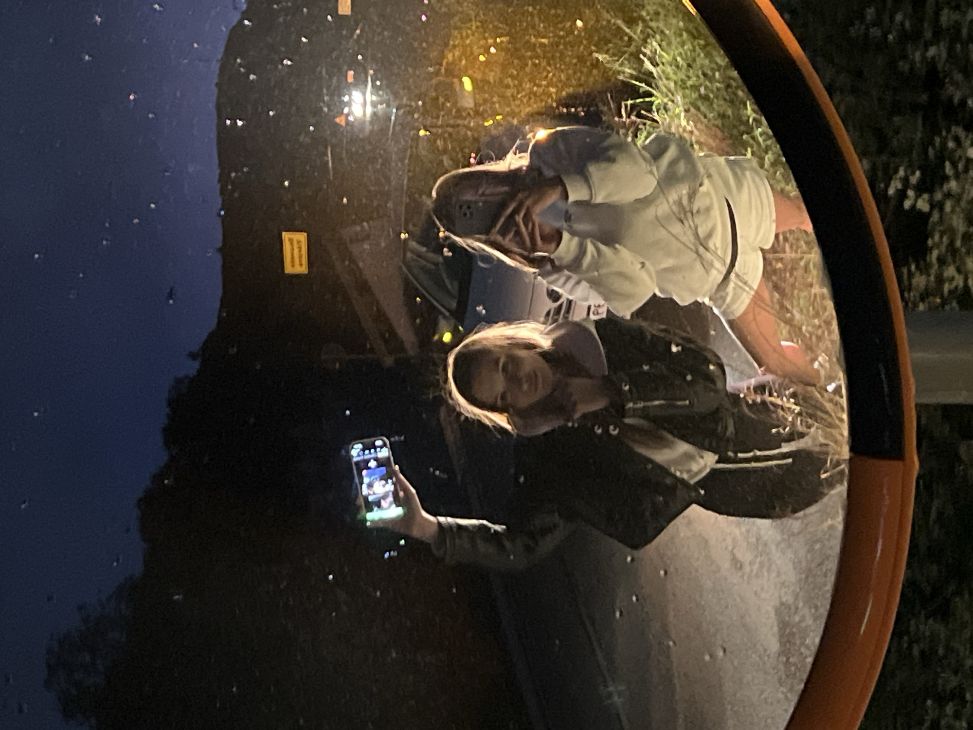 This media file captures a mirror selfie of two young women taken at night in Visnjeva, Montenegro. The image is a reflection in a large, round convex safety mirror, distinguished by its orange rim.
In the foreground of the reflection, a woman with light brown hair and a slight smile holds a smartphone, seemingly capturing the image. She is wearing a dark jacket over a lighter top. Her phone screen is visibly illuminated, displaying an app interface. Behind her, another woman is partially visible, wearing a light-colored hooded sweatshirt and shorts. Her face is partly obscured by her hands, suggesting a playful or shy pose, and she appears to be smiling.
The activity taking place is the two women posing together for a selfie or video, utilizing the unique distortion of the convex mirror.
Notable details indicate it is nighttime, with the scene illuminated by artificial light sources, likely streetlights, reflecting in the mirror. The mirror's surface is covered with numerous small, bright specks, suggesting it might be wet from rain or mist. Reflected in the mirror are elements of an outdoor setting: what appears to be a road surface, parts of a vehicle, and dark vegetation or bushes along the roadside. A small, rectangular yellow sign with indecipherable text is also reflected in the upper left area of the mirror. The overall mood appears casual and joyful.